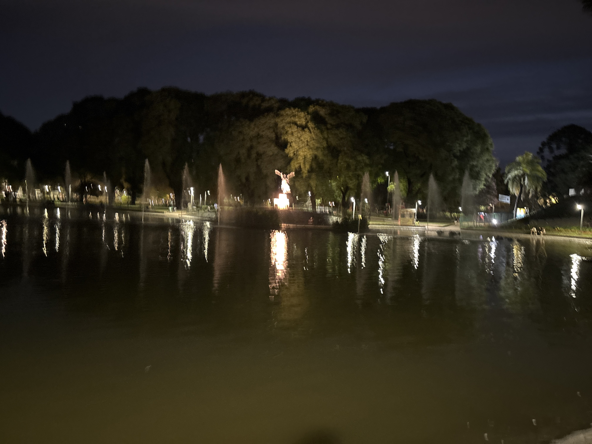 The image shows a lake at night in Buenos Aires, Argentina. The lake reflects the lights from the surrounding trees and a statue in the middle of the lake. Fountains are visible along the edge of the lake. The statue is illuminated and has a figure with outstretched arms. Trees line the bank of the lake. Lampposts are scattered throughout the area. Some people are walking along the bank in the distance.