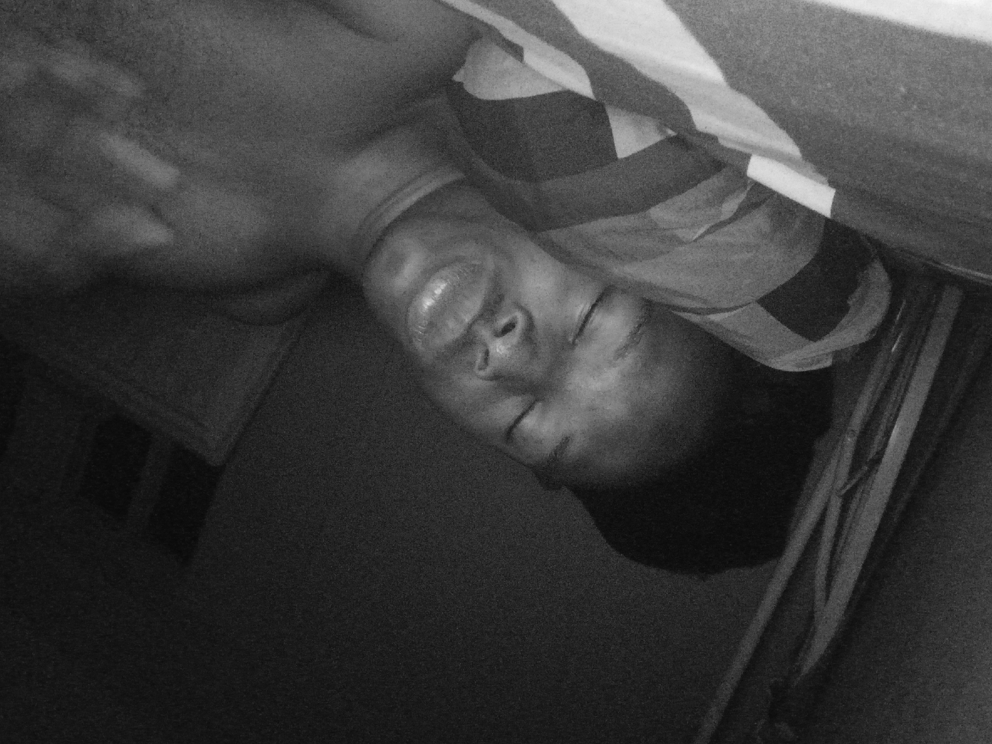 A monochrome, low-angle photograph shows a person lying down, seemingly in bed. The person is Black, and has their eyes closed. A striped sheet or blanket partially covers their head. The setting appears to be indoors, with part of a wall visible in the background and what appears to be a window. Due to the quality and lack of context, specifics related to the city of Lagos, Nigeria are unidentifiable.