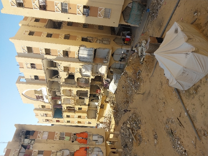 This image depicts a severely damaged urban residential area in Khan Yunis, Palestinian Territory, under a clear, light blue sky. Three multi-story, light-tan residential buildings, each featuring decorative architectural elements like arched windows and patterned railings, exhibit extensive structural damage. Walls are significantly breached or missing, exposing interior rooms filled with debris and collapsed elements. Numerous windows are shattered or entirely absent, and facades bear visible impact marks and pockmarks. The central building shows particularly severe damage, with multiple floors largely open to the elements and its distinctive arched facade at the top center partially destroyed. The leftmost building has large sections of its exterior stripped away, revealing bright orange fabric or tarps covering some openings. Laundry hangs from various damaged sections of the buildings, particularly at lower levels of the central and right structures.
The foreground and midground consist of a large, uneven expanse of sandy ground heavily littered with concrete rubble, fragmented building materials, and twisted metal debris. Several dark pipes or conduits run horizontally across this sandy area. In the lower right foreground, a single, soiled white canvas tent with a peaked roof is erected on the sand.
A lone individual, dressed in dark clothing, walks across the sandy debris field in the midground, moving towards the left. Near the base of the central building, to the right, a small group of approximately 3-4 individuals, including adults, are visible standing or moving amidst the rubble. Further indistinct figures are discernible under an archway at the base of the leftmost building. The overall scene conveys widespread destruction with ongoing human presence amidst the ruins.