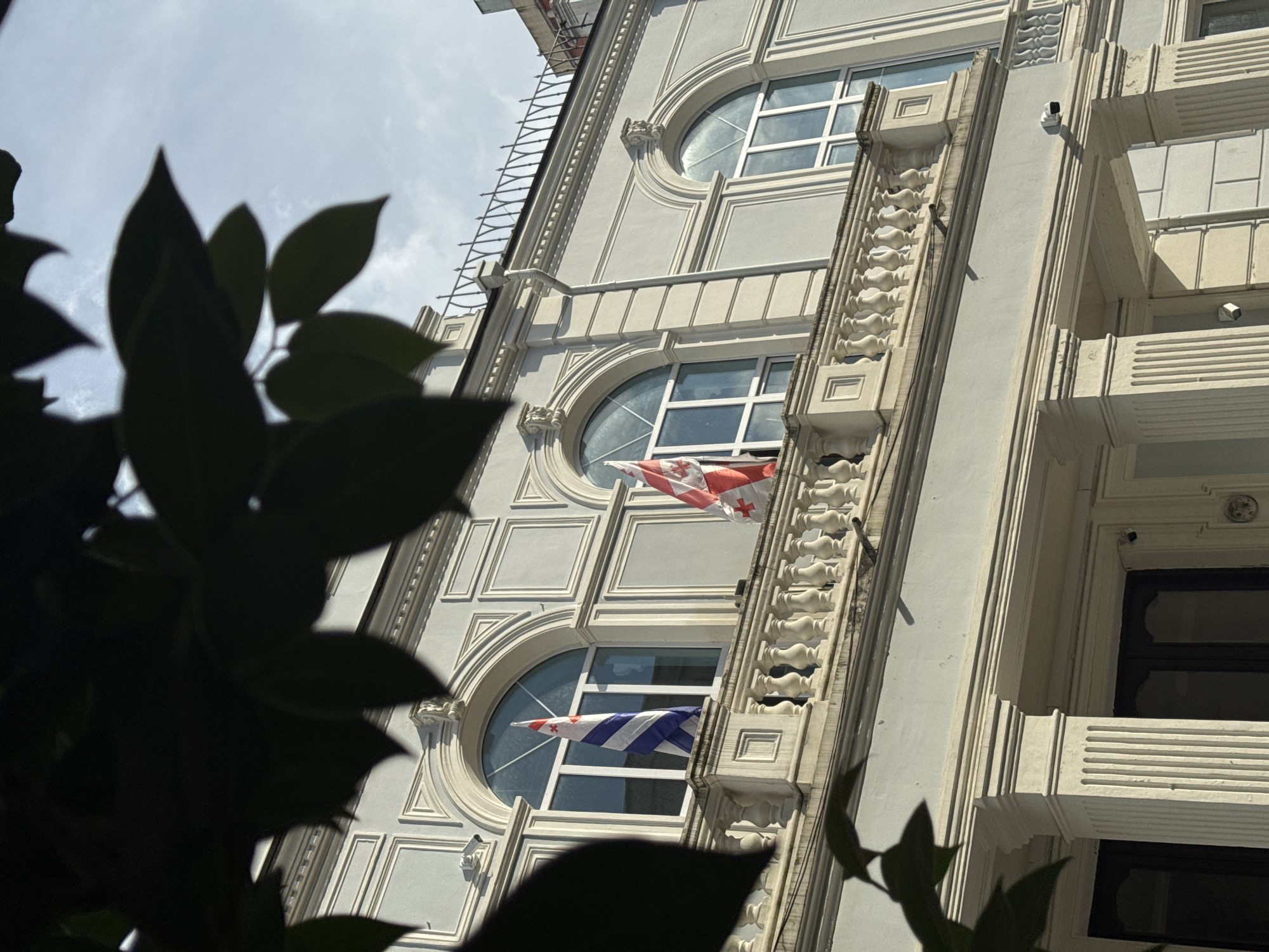 The image is a close-up, low-angle shot of a building facade in Batumi, Georgia, partially obscured by dark green foliage in the foreground. The building appears to be an older, ornate structure with light-colored stone or stucco. It features multiple levels with large, arched windows. Two of these windows are clearly visible, each containing a flag. The upper window displays a flag with a red cross on a white background, and the lower window shows a flag with blue and white stripes and a red cross in the upper hoist quarter. The flags are unfurled and appear to be waving slightly.
Architectural details of the building include decorative moldings, fluted columns, and what appear to be balconies with balustrades. The windows themselves have multiple panes. The sky above is overcast with some light clouds, suggesting it is daytime, possibly late morning or afternoon. The overall impression is one of historical architecture and a nationalistic display. No people are visible in the image. The scene is static, with the flags being the only elements in motion. The lighting suggests it is a bright, possibly sunny day despite the overcast sky.
