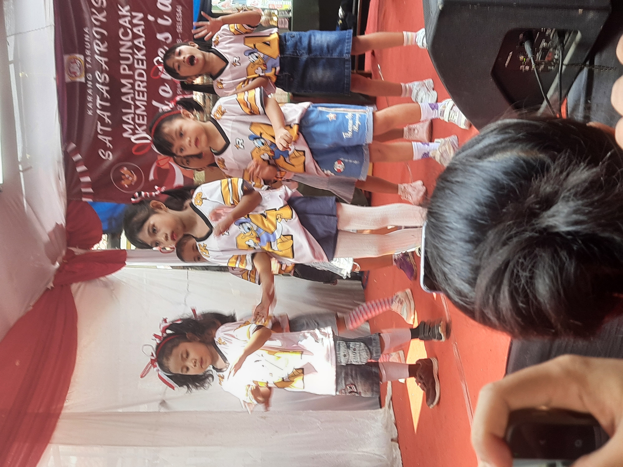 In the city of Bandung, Indonesia, a group of young girls in matching shirts and skirts are performing on a red stage, adorned with red and white banners. The banners, featuring the text "KARANG TARUNA SATATAS ARIKS MALAM PUNCAK KEMERDEKAAN INDONESIA" and a depiction of the Indonesian flag, suggest the event is a celebration of Indonesian Independence Day. The girls are wearing white t-shirts with cartoon characters and the number "21" printed on them, some with red bows in their hair. They are likely performing a song or dance. One girl in the foreground is holding her hand up, as if to clap or wave. The background reveals a white curtain and more red and white fabric draped from above. The overall scene captures a lively and festive atmosphere, typical of a community event celebrating national pride.