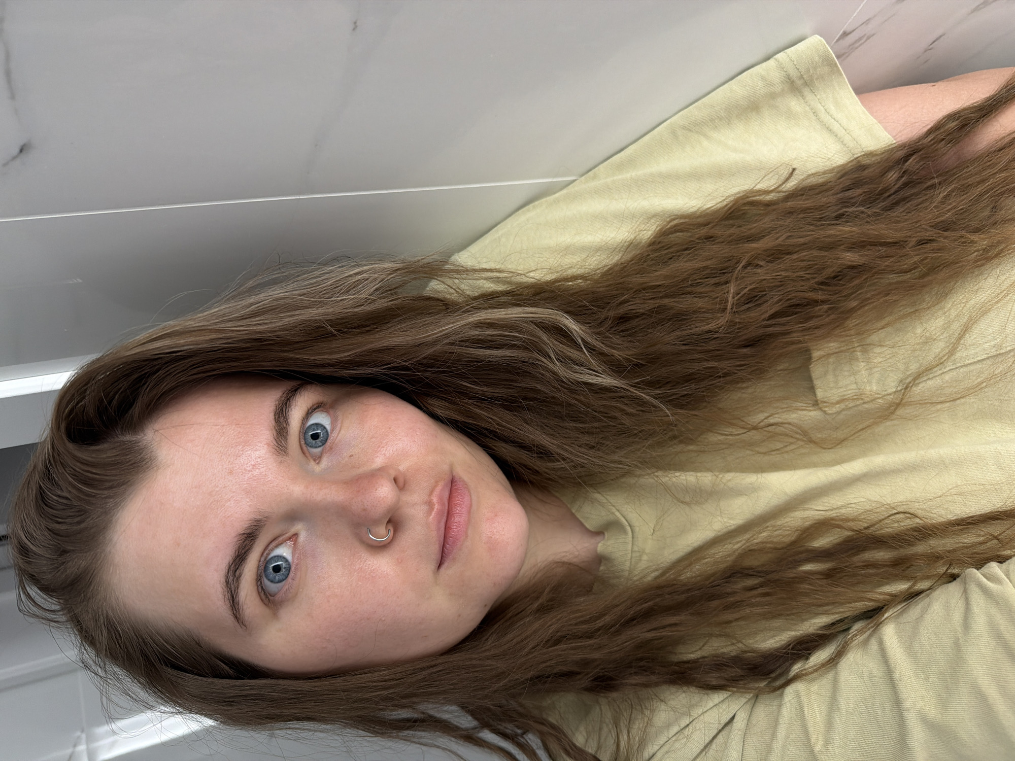 A light-skinned individual with blue eyes and brown hair is pictured indoors. The person is wearing a light yellowish-green shirt. A silver ring is visible in their left nostril. The background consists of white tiled walls. There is no information indicating this image was taken in Chaikovsky, Russia.