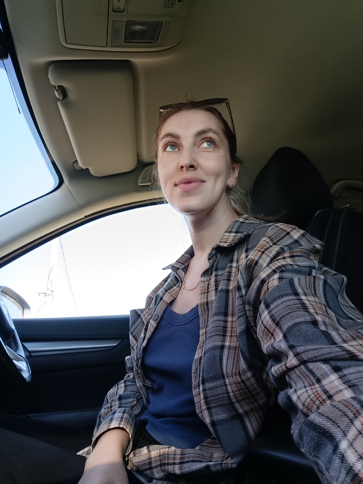 A young woman sits inside a car, possibly a compact SUV, in an indeterminate location. She is wearing a navy blue t-shirt and a gray and brown plaid flannel shirt. She has light skin and light brown hair, with sunglasses resting on top of her head. She is looking upward and to the left with a slight smile. The car's interior is a combination of gray and black. A sun visor is lowered on the driver's side. The steering wheel and dashboard are visible. The seat behind her is black. It's daytime with bright sunlight visible through the windshield. The location in Chaikovsky, Russia is unknown.