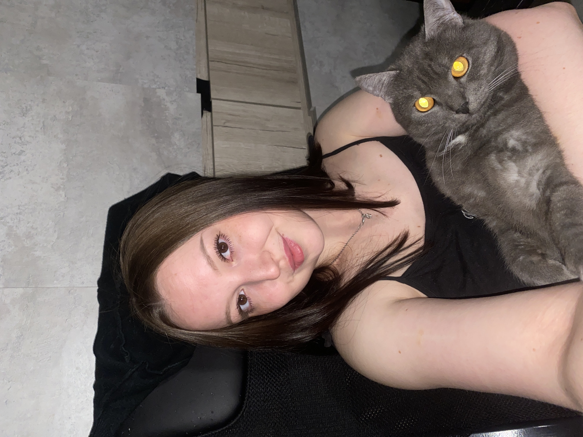 A woman and a gray cat are visible in the image. The woman, with brown hair and fair skin, is wearing a black tank top and a silver necklace. She is reclining on a black surface, possibly a chair or cushion. Her face is turned towards the camera with a slight smile. The cat is resting on the woman's arm, and it has golden eyes that are prominently illuminated. The background includes a light gray wall and a light brown wooden cabinet. It is not possible to verify location.