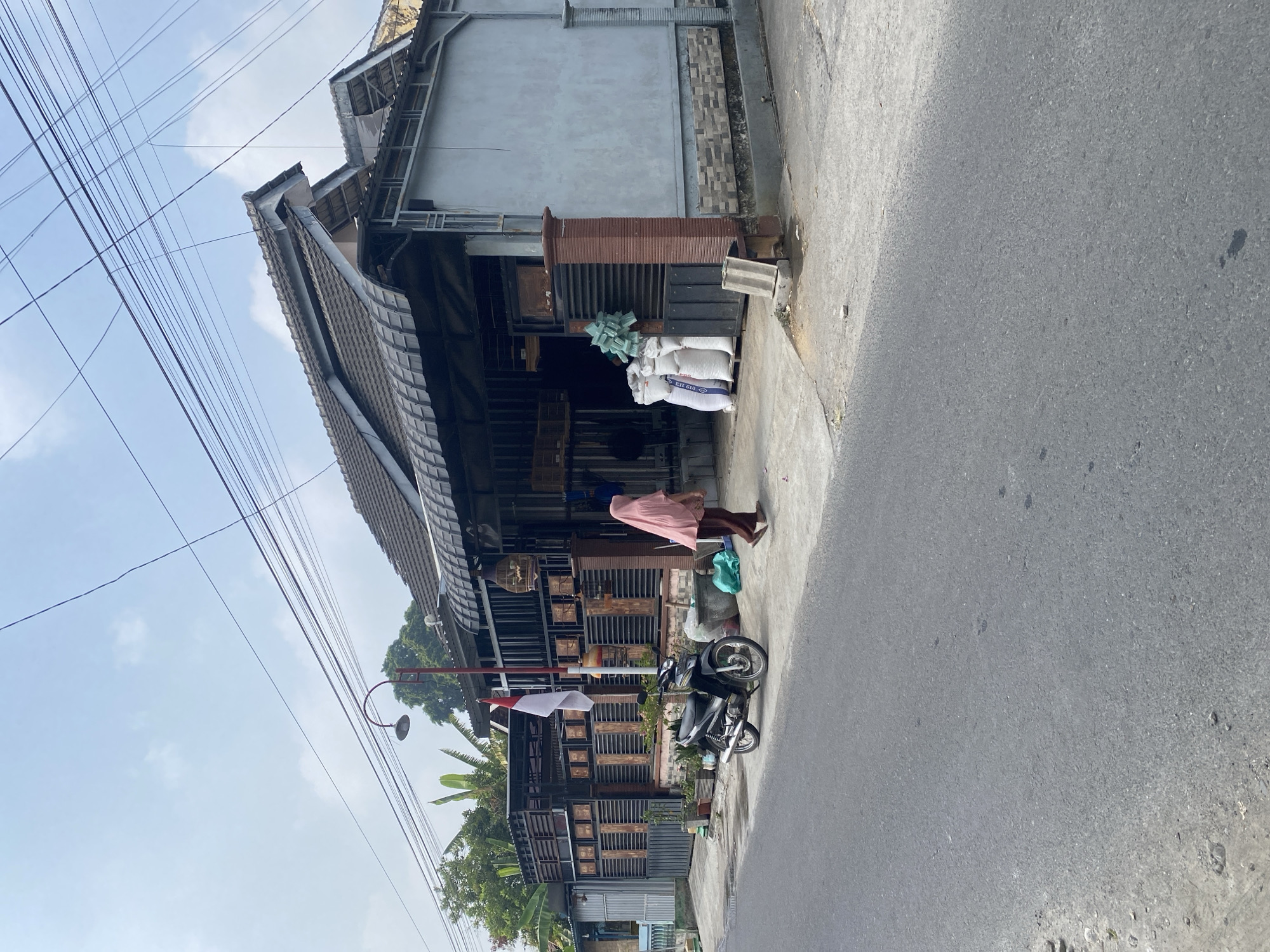 The image depicts a street scene in Kraton, Indonesia, on a clear day. In the foreground, a gray asphalt road runs from left to right. A concrete sidewalk runs parallel to the road. On the sidewalk, a black motorcycle is parked. To the right of the motorcycle, a person wearing a pink hijab and brown clothing walks away from the camera. Behind the person and motorcycle, there is a building with a dark-tiled roof and an open-fronted section that appears to be a shop or workshop. Several white sacks are stacked in front of the shop. A red and white Indonesian flag is displayed on a pole near the motorcycle. The building is flanked by fences and other structures. Overhead, a network of dark electrical wires crisscrosses the sky. The environment appears to be a residential or commercial area in a town or city.