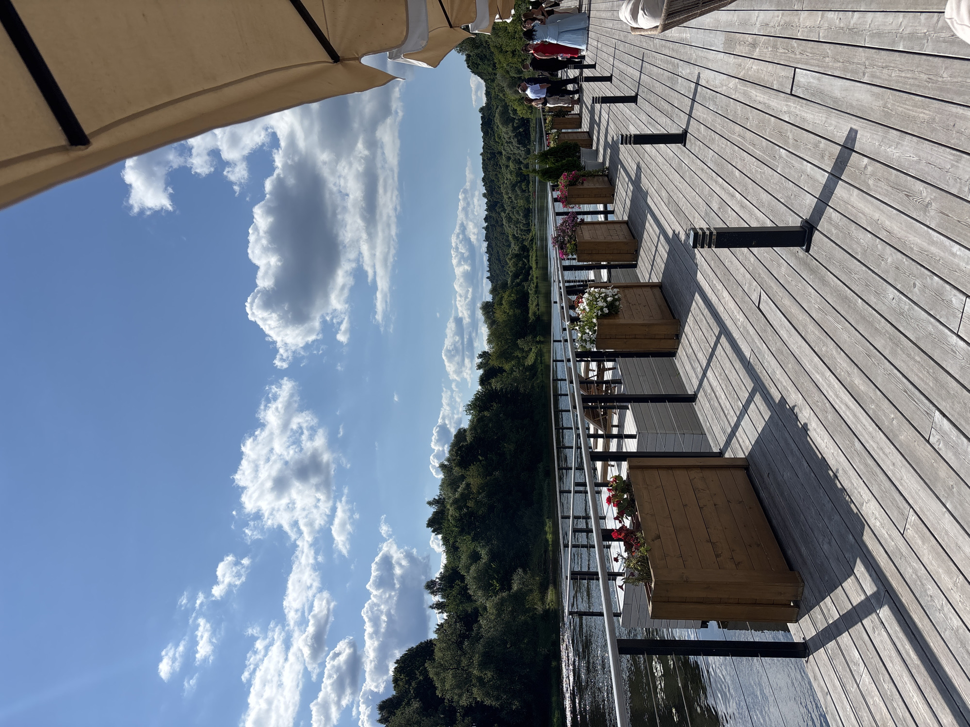 A wide-angle outdoor shot captures a wooden deck extending over the water, likely a river or lake, under a partly cloudy blue sky. The deck is adorned with wooden planters filled with flowers and several modern black bollard lights spaced along its edge. In the background, a lush green forest borders the water, with a distant view of rolling hills. A group of people, dressed in formal attire, are gathered on the right side of the deck, appearing to be engaged in conversation. The scene suggests a leisurely gathering or event taking place at a waterfront venue. This location is situated in Zvenigorod, Russia.