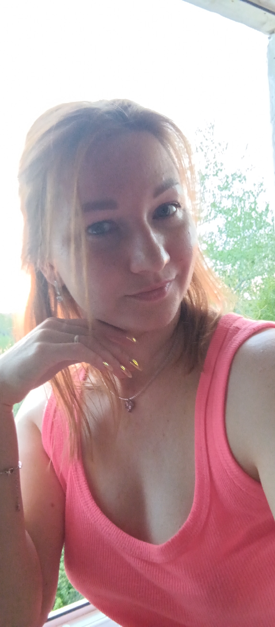 A close-up shot depicts a woman with light skin and reddish-blonde hair, wearing a bright pink tank top. Her left hand is raised, with her fingers touching her neck. She has a ring on her finger and a necklace with a pendant is visible around her neck. Her nails are long and appear to be painted gold. The background is blurry, showing foliage and what appears to be a window frame. The location is identified as the city of Unknown, Unknown.