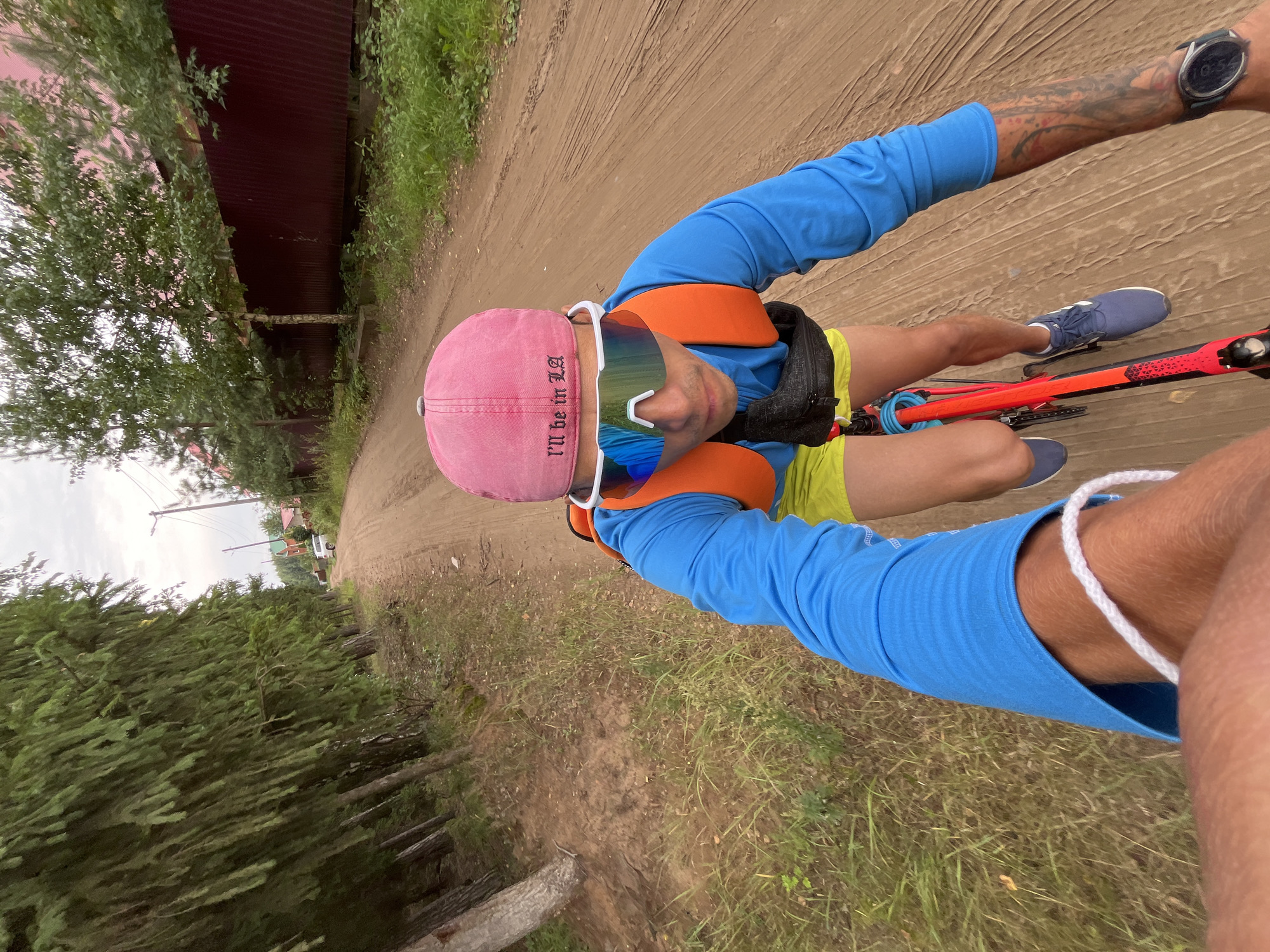 A first-person perspective shot shows a person riding a bicycle on a dirt road lined with trees and houses. The rider wears a blue long-sleeved shirt, yellow shorts, a pink cap with the text "I'll be in LA" embroidered on it, and white sunglasses. An orange vest is worn over the shirt, and a black fanny pack is visible around the waist. The rider's right arm is visible, showing a tattooed sleeve and a black smartwatch. The road is dusty and appears to be in a rural or suburban area. The background includes houses with red roofs and green trees, characteristic of a Russian dacha or village setting. The sky is overcast. The location is identified as Chaikovsky, Russia, likely a dacha community or rural road on the outskirts of the city.