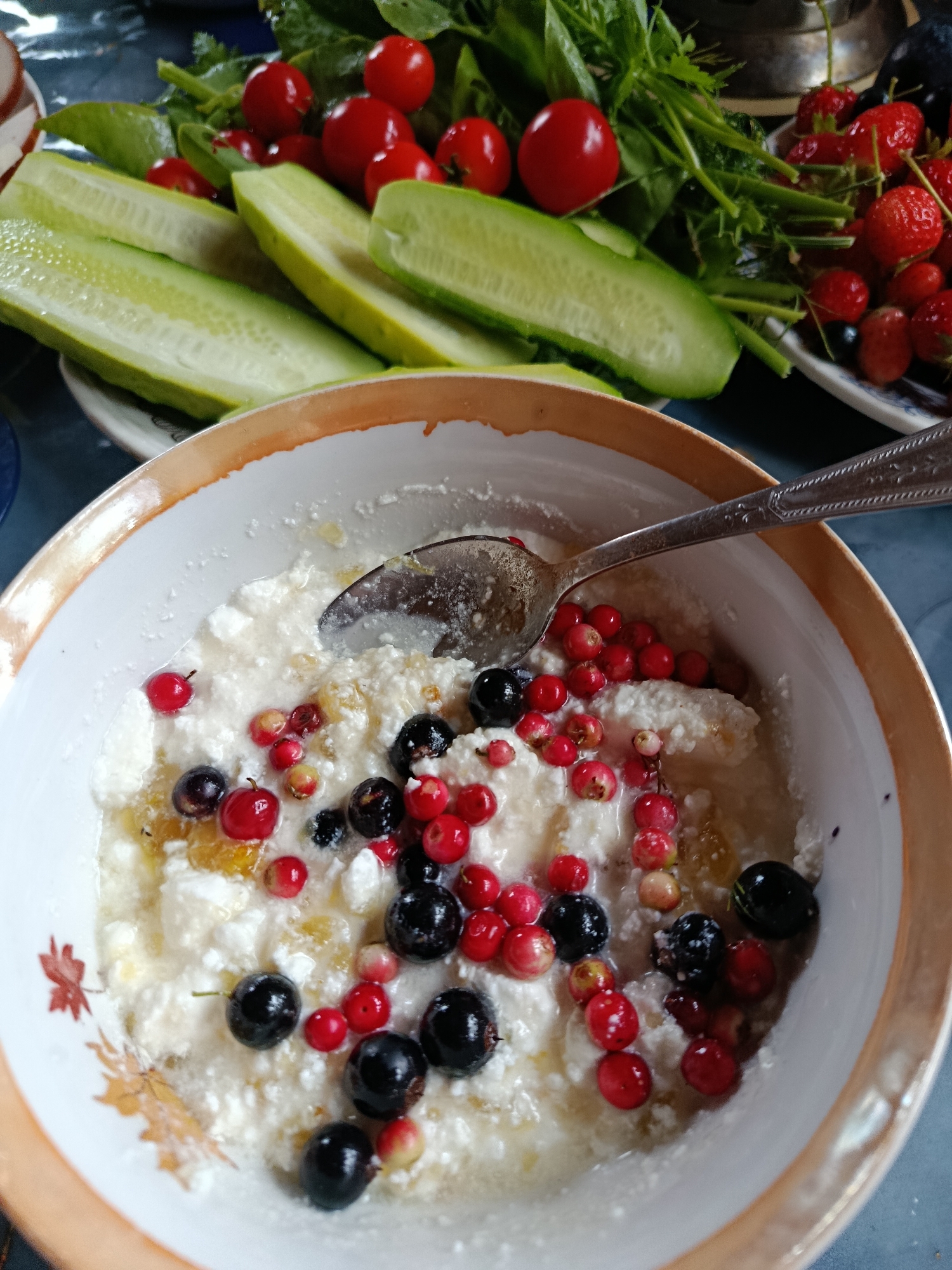 A bowl of cottage cheese topped with red and black berries and a spoon is the main subject in this close-up, eye-level shot. The bowl has a light brown rim with a faded floral pattern. In the background, sliced cucumbers, cherry tomatoes, and fresh greens are arranged on plates. The scene appears to be indoors, possibly on a table. This image was taken in the city of Vereya, Russia.