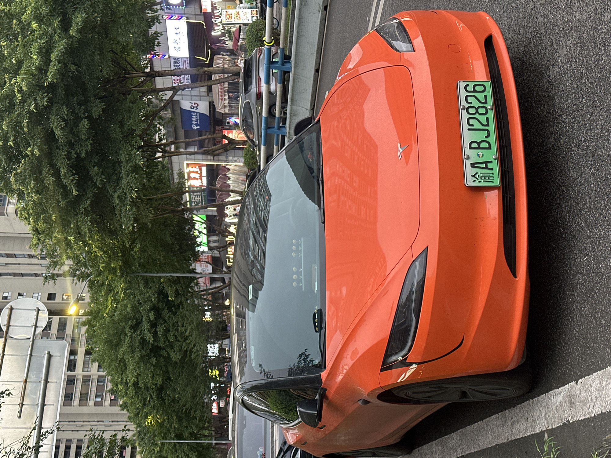 An orange electric car, identified by its Tesla logo and license plate "渝A BJ2826", is parked on a street in the Chongqing High-tech Zone, China. The car is a prominent subject, viewed from its front-right quarter. Behind the car, a row of trees with lush green foliage lines the street, partially obscuring buildings in the background. These buildings appear to be multi-story structures with various illuminated signs indicating businesses. The scene suggests an urban environment during either late afternoon or early evening, as indicated by the artificial lighting from the signs and streetlights starting to glow, and the soft, diffused natural light. The weather appears to be calm, with no strong indications of wind or rain. The overall atmosphere is that of a typical city street, with elements of commerce and transportation. The image captures a slice of everyday life in a developed urban area in China.