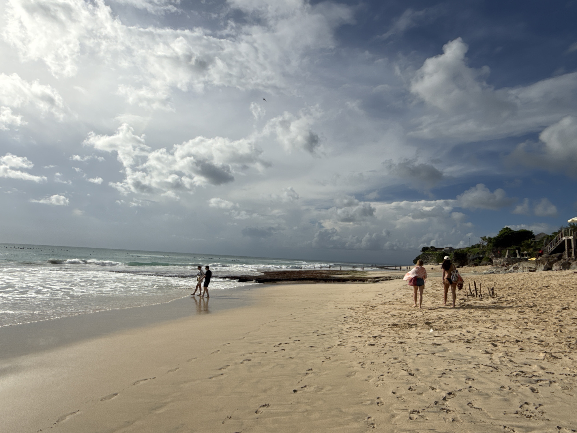 A panoramic view captures a lively beach scene under a cloudy sky. The ocean, with gentle waves breaking into white foam, stretches across the left side of the frame. In the distance, several small figures, likely surfers, can be seen in the water.
The sandy beach, wet near the waterline and drier further back, dominates the lower half of the image. Numerous footprints are visible in the sand, indicating recent activity.
Several people are present on the beach. Towards the left, two individuals—a male in dark clothing and a female in lighter attire—walk together along the water's edge. Further to the right and slightly closer to the foreground, two other females walk away from the camera. One wears a dark bikini and a light-colored hat, carrying a bag, while the other wears shorts and a patterned shawl.
The sky is filled with a mix of bright white and grey clouds, suggesting an overcast day with the sun partially obscured but still casting a luminous glow, particularly on the left side. Patches of blue sky are visible between the clouds. A small, dark object, possibly a bird or a kite, is discernible against the clouds in the upper-middle section.
On the far right, the beach transitions into a rocky area, beyond which a vegetated hillside rises, dotted with buildings or structures, and a wooden staircase descending towards the sand. Some wooden stakes or posts are embedded in the sand near the rightmost walkers.
The scene depicts a relaxed daytime atmosphere at a tropical beach, consistent with the Kuta Selatan, Indonesia context. There is no visible text in the image.