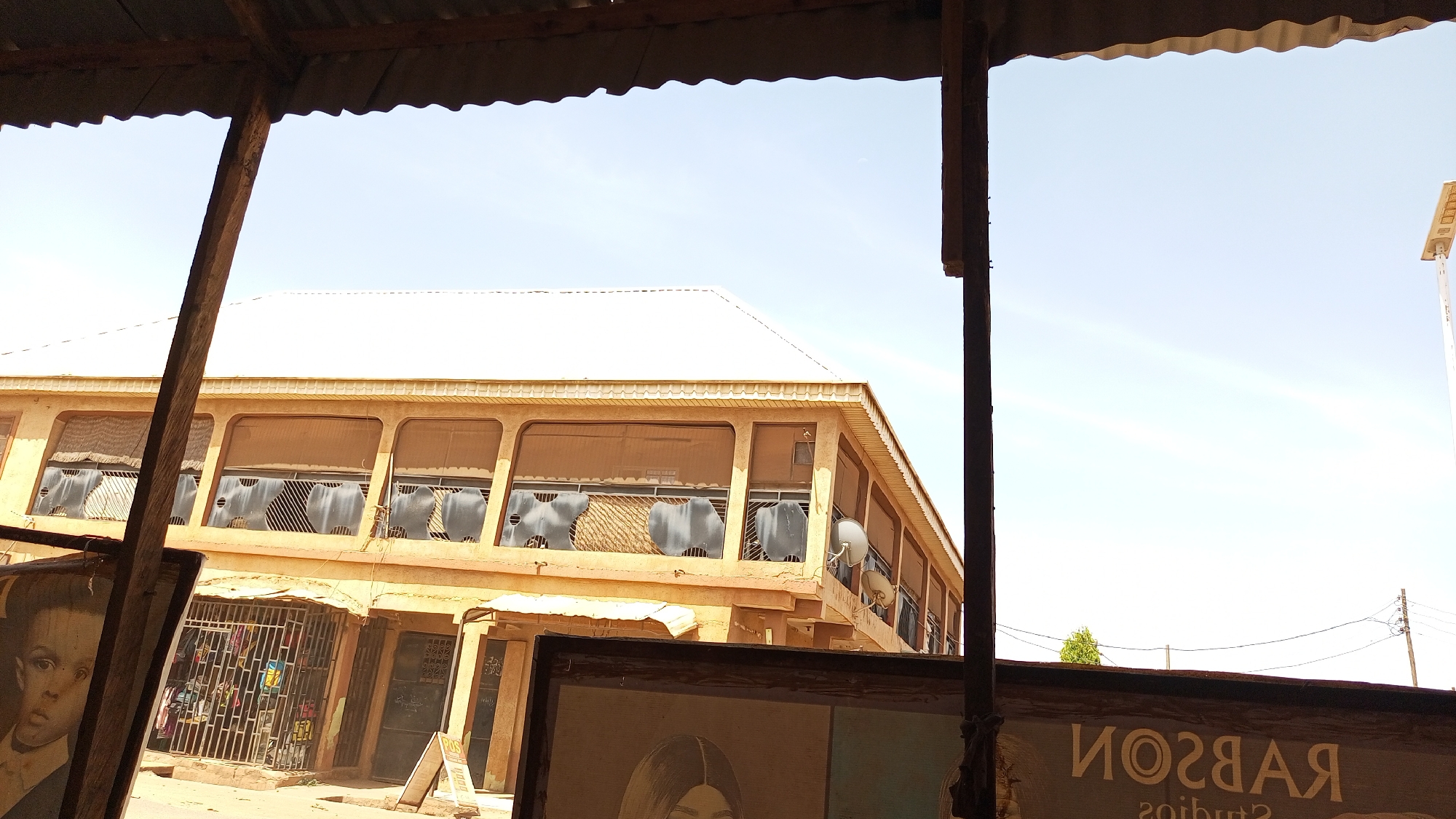 The image captures a street scene viewed from under a corrugated metal roof, with dark wooden support beams prominently in the foreground. The vantage point looks out towards a two-story building under a clear, bright blue sky.
In the immediate foreground, part of a dark frame or sign is visible on the left, displaying a monochrome portrait of a young child's face. On the bottom right, another large, dark rectangular sign or frame is visible, which appears to be a mirrored reflection, showing the text "ЯOSЯAЯ" (RABSON) and "2oibut2" (Studios) beneath it.
The main subject is a large, yellowish-brown building with a light-colored, possibly white or silver, gabled roof. The upper floor features a series of open, arched or rectangular balcony sections. Behind these openings, several gray objects, possibly chairs, seating units, or ventilation components, are visible. Multiple small satellite dishes are mounted on the side of the building towards the right. The ground floor of the building consists of several commercial establishments or shops, protected by metal security grates. One shop, visible on the left, appears to sell clothing, and an A-frame sign stands outside one of the ground-floor units.
The background is dominated by a bright, clear blue sky, suggesting fair weather. On the far right, a modern street light or utility pole equipped with solar panels is visible against the sky. A small green bush or tree can be seen in the distance above the mirrored sign.
The overall impression is that of a bustling commercial area during a bright day, possibly in an urban or semi-urban setting, likely in a warm climate. No specific activity or event is taking place; the image is a static snapshot of the environment.