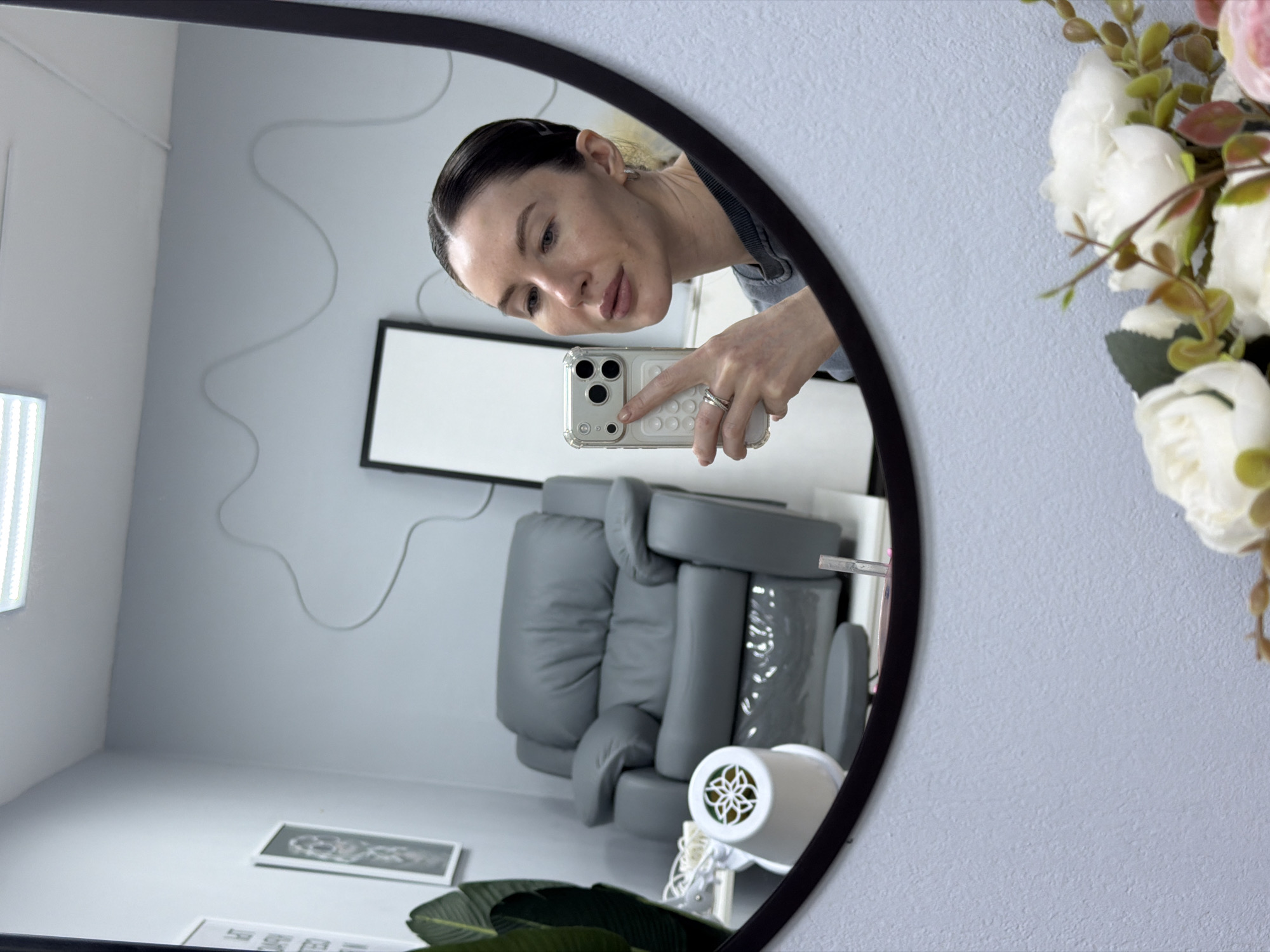 The image displays a mirror selfie taken by a woman in what appears to be a beauty salon or spa. The woman is reflected in a black-framed oval mirror, holding a light-colored iPhone in a clear, textured case with her right hand, which wears several rings. She has dark hair pulled back, fair skin, and is wearing a grey top, with a neutral expression as she focuses on the camera.
The room reflected in the mirror features light grey walls and flooring. A prominent grey, segmented chair or treatment bed takes up much of the reflected space. On the floor, a white decorative air purifier or fan with an ornate circular grill is visible. A winding grey cable is affixed to the wall, and a black-framed object with a white screen is mounted on the wall. A vertical strip light on the left side of the reflection illuminates the area. In the lower foreground of the reflection, a plant with large green leaves is partially visible, alongside two small framed pictures on the floor.
Outside the mirror's frame, on a light blue textured wall, a bouquet of artificial flowers with white and pink roses and green foliage is positioned to the right. No visible text is discernible in the image.