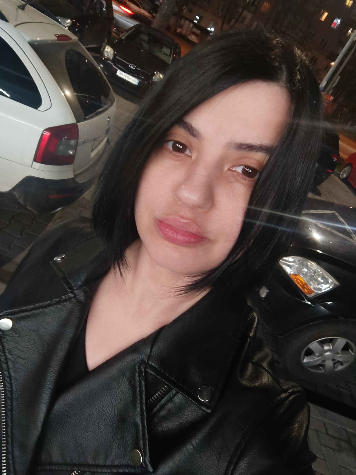 A medium shot depicts a woman with dark hair and a black leather jacket, likely taken at night in an urban environment. The woman's face is centrally framed, revealing soft features, and her gaze is directed slightly off-center. Her hair is styled in a sleek, short bob. She wears a black leather jacket, partially unzipped, revealing a black top underneath. In the background, blurred vehicles suggest she is near a street or parking area. To her left, a white car is partially visible, and a darker car is positioned behind it. The lighting seems to be from street lamps or other artificial sources, casting shadows and highlights on her face and jacket. The location is suggested to be the city of Unknown, Unknown.