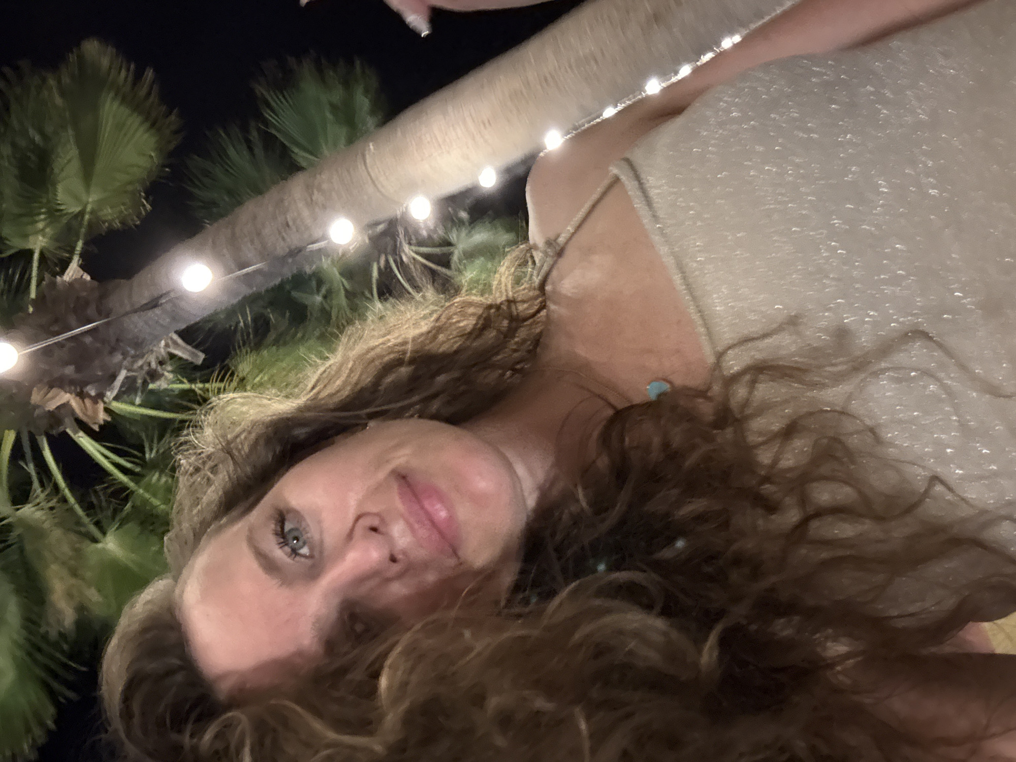 A woman is taking a selfie at night. She has long, wavy brown hair and blue eyes. She is wearing a sparkly, light-colored top. In the background, there is a palm tree with string lights wrapped around it. The location is not specified in the image.