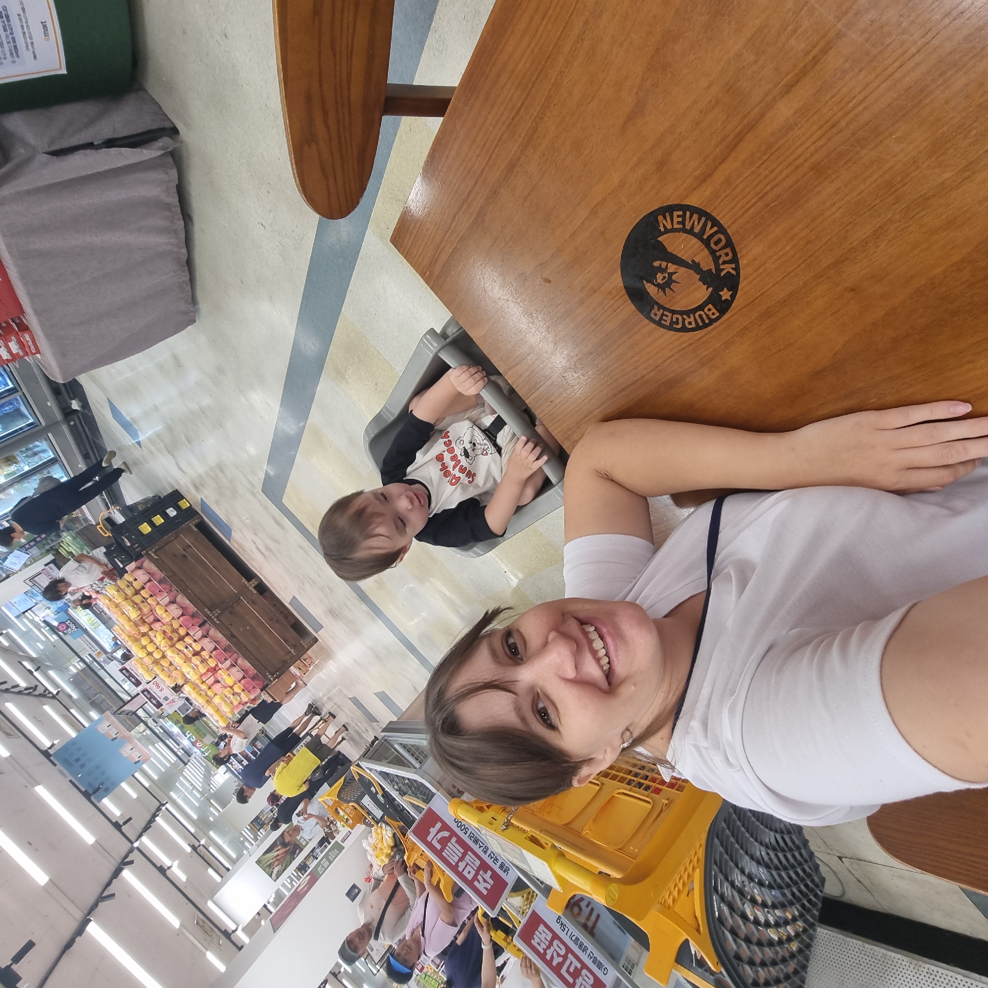 In a brightly lit supermarket in Wonju-si, South Korea, a woman in a white t-shirt smiles at the camera, her left arm resting on a wooden table. To her right, a toddler sits in a high chair, wearing a black and white shirt with a graphic. The table has a circular black logo with the text "BURGER NEW YORK". In the background, other shoppers and grocery carts are visible, with displays of produce and shelves stocked with goods. The floor has a patterned tile design, and the ceiling is lined with fluorescent lights.