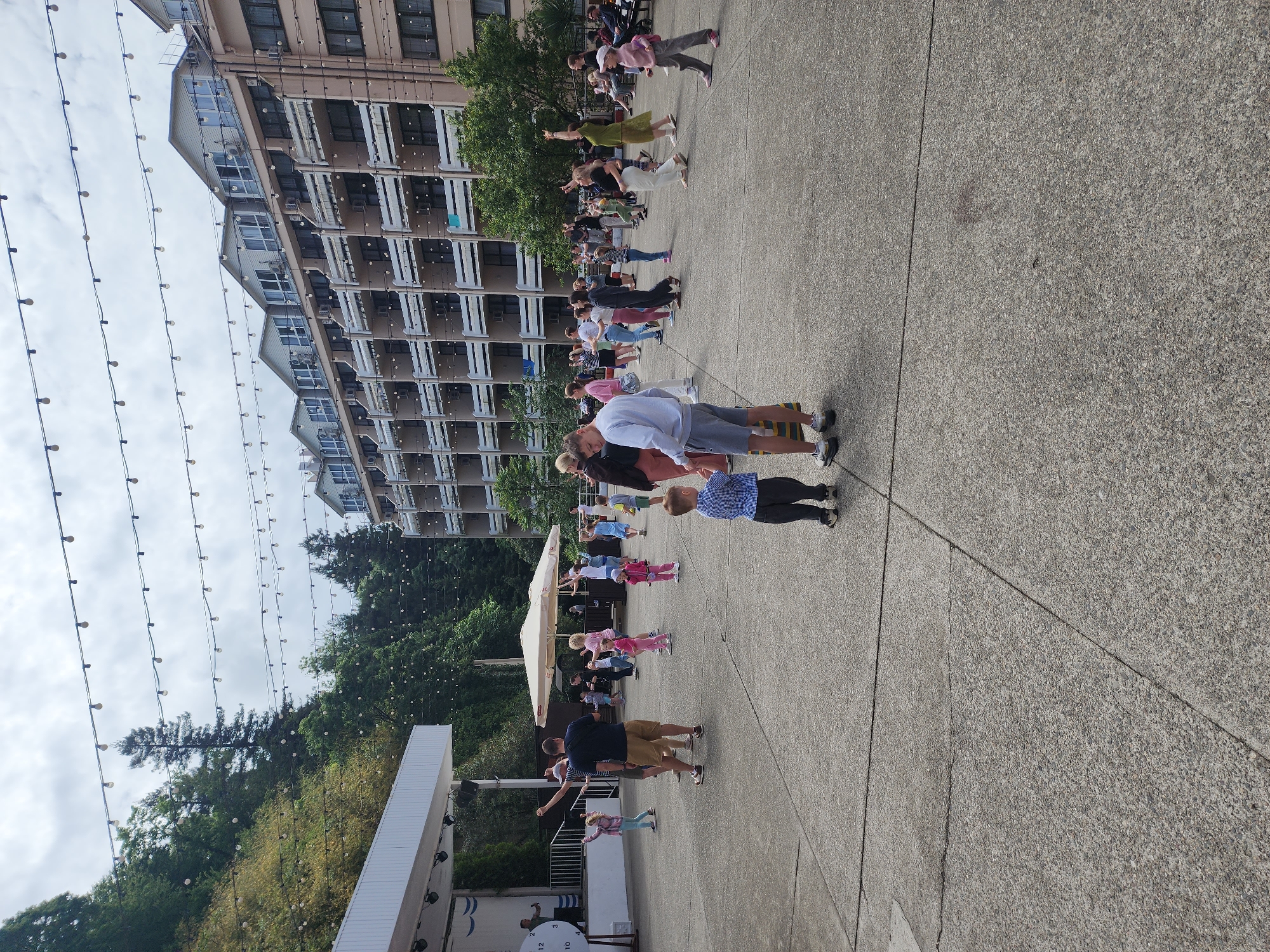 The image shows a group of people, mainly adults and children, doing a group exercise or dance outdoors, likely in a public square or plaza. They are aligned in rows, with their arms extended to the side. A stage with some branding or design is visible in the lower left of the image. In the background is a multi-story building and a hillside covered with trees. Overhead, strings of lights are hung across the area. The location is not specified, but is potentially in Sochi, Russia.