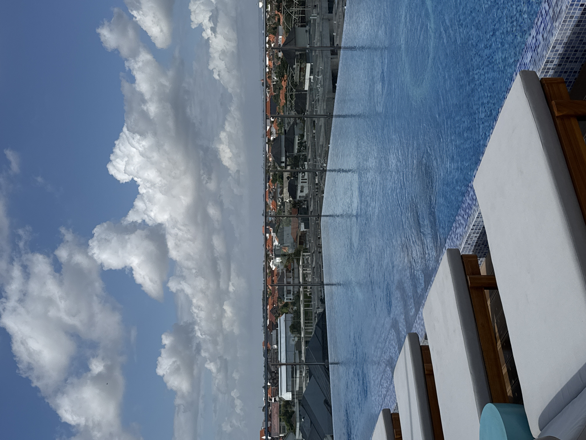 An outdoor infinity pool overlooks a dense urban landscape in North Kuta, Indonesia. The pool, filled with clear blue water, is lined with white chaise lounges and a small blue side table. Beyond the pool, a glass railing separates the pool area from the city. The city below is characterized by a multitude of buildings with red-tiled roofs, interspersed with some modern structures. In the background, the sky is a vibrant blue, adorned with large, fluffy white clouds. The overall scene captures a moment of relaxation and urban vista.