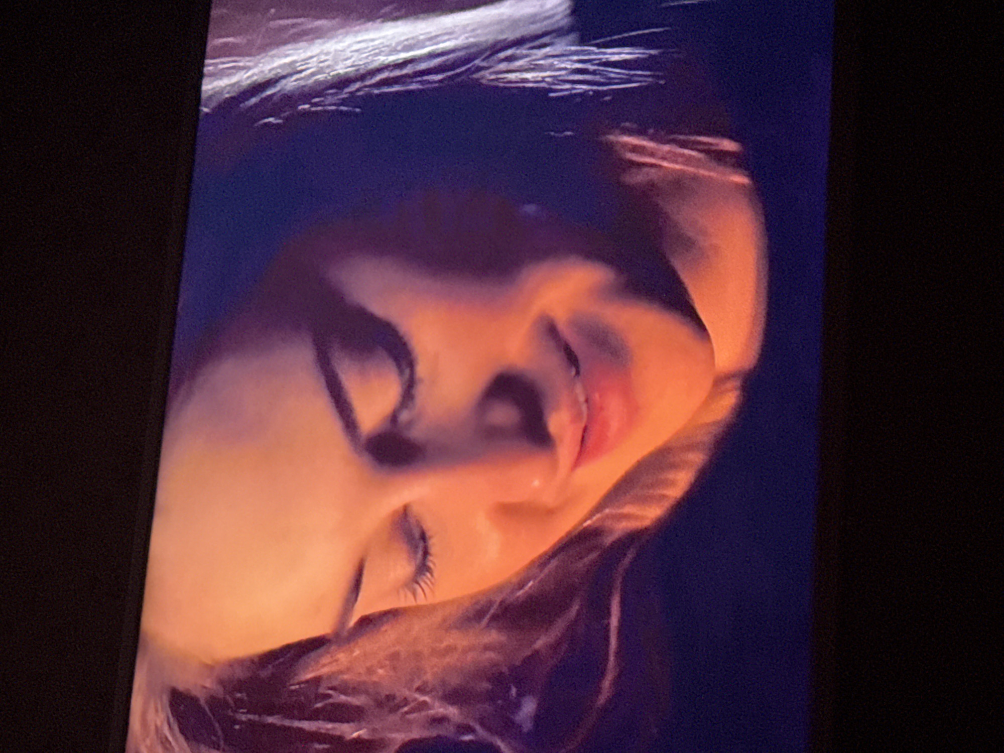 A close-up, tilted image depicts a person's face. The person has closed eyes and a slight smile. The skin tone is warm, possibly due to the lighting. Dark hair is visible at the top and bottom of the frame. The background is dark and indistinct. There is no information to suggest the image was taken in Mytishchi, Russia, or any other specific location.