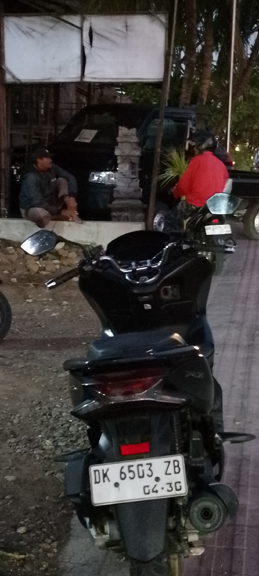 In Pekutatan, Indonesia, an outdoor scene at dusk captures two individuals and several vehicles. In the foreground, the rear of a black scooter is prominently displayed, showcasing its license plate which reads "DK 6503 ZB" and "04.3G". The scooter has a sleek design with a visible exhaust pipe and a red reflector above the license plate. To the left of the scooter, a black motorcycle mirror is angled towards the viewer.
Further back, a man wearing a dark jacket and cap is seated on a concrete ledge, looking towards the left. Beside him, a black vehicle, possibly a van or truck, is parked. Behind this vehicle, another person is seated on a scooter, wearing a bright red jacket and a helmet, facing away from the camera. This individual is positioned near a larger dark-colored vehicle. The environment appears to be a roadside or a public area, with a mixture of gravel and paved ground. Trees are visible in the background, suggesting a tropical setting. A white tarp stretched over a frame is also visible in the upper left portion of the image. The lighting suggests it is either early morning or late evening.