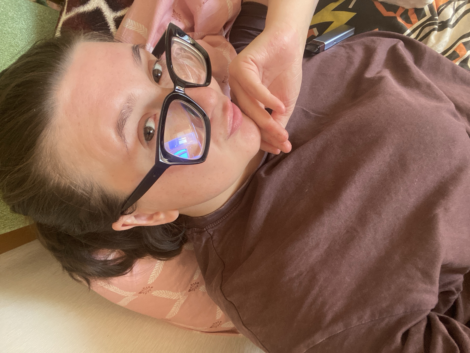 A woman with brown hair is lying down, wearing glasses with black frames and a brown t-shirt. Her right hand is near her face. A silver electronic device is visible near her right shoulder. She's resting on a patterned pink pillow, which itself is lying on a white surface. A colorful blanket or pillow is in the background. There is no definitive information to indicate this photograph was taken in Chaikovsky, Russia.