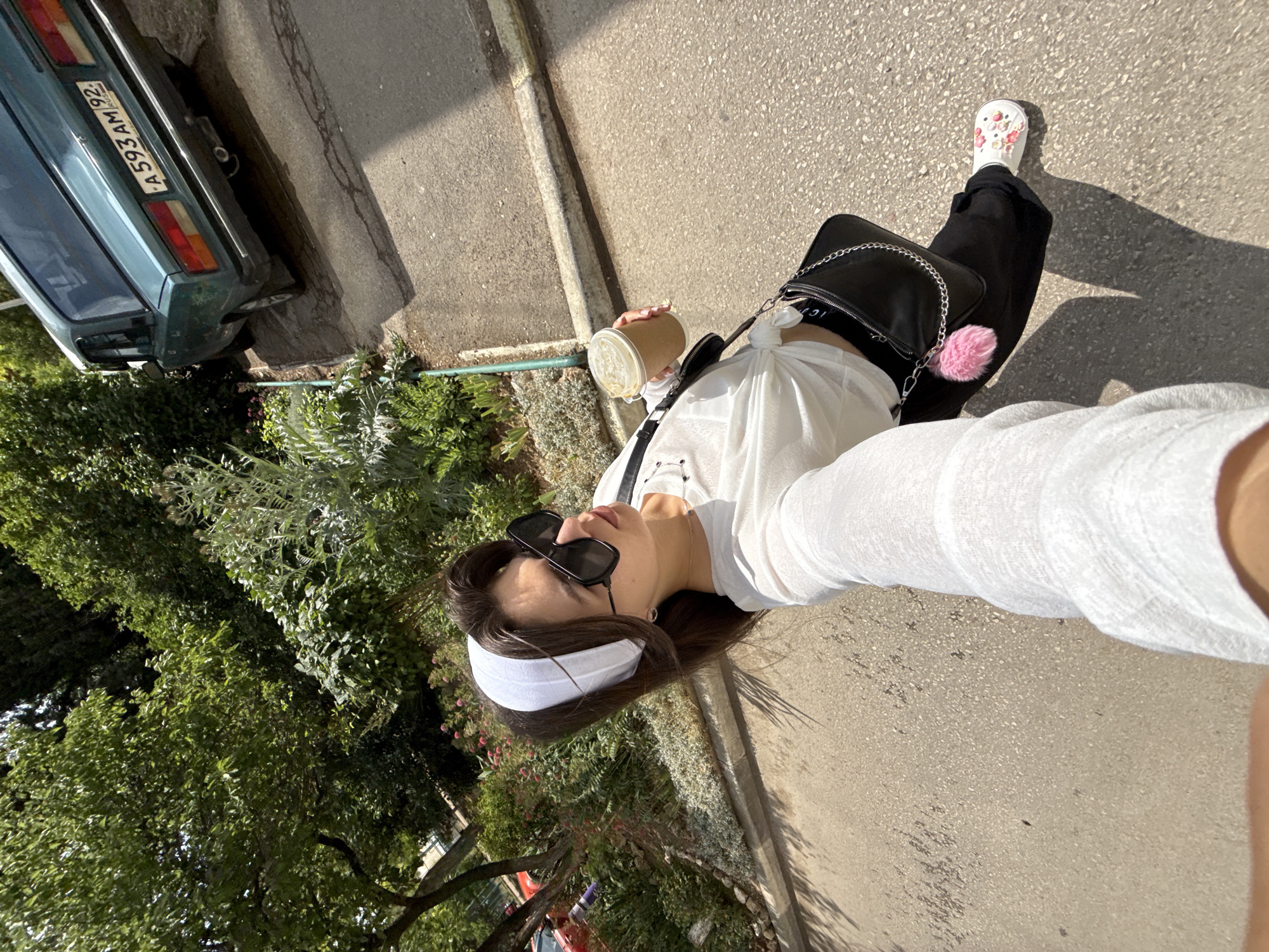 A young woman with a white headband, black sunglasses, and a white long-sleeved top is walking on a paved street. She is holding a disposable coffee cup in her right hand and has a black shoulder bag with a silver chain and a pink pom-pom attached to her left side. She is wearing black pants and white clogs decorated with pink flowers. In the background, there is a turquoise car with a license plate that reads "A593AM92" parked on an incline. Lush green trees and plants are visible on the left and behind the car. The sun is casting shadows on the street.