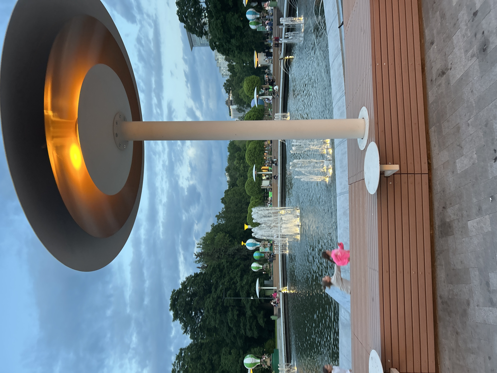 The image depicts an urban park or plaza scene during dusk or early evening. In the top left, a large, modern light fixture with a golden-orange inner surface and a white exterior is prominent, casting a warm glow. It is mounted on a white pole.
The main part of the scene features a long, rectangular water feature, possibly a canal or large fountain, running horizontally across the middle of the frame. Multiple illuminated water jets create fountains within the water. Alongside the water feature, there's an expansive wooden deck made of parallel planks, providing a pedestrian area. Several white, circular platforms or tables are positioned on this deck.
Scattered along the edge of the water and the deck are decorative elements resembling hot air balloons, primarily in light green and blue/white colors, some of which appear to be lit from within. Various people are visible, some standing or walking on the deck. One person in a bright pink top is distinct on the right side of the wooden area.
In the background, a lush green treeline borders the park, with some tall buildings partially visible behind the foliage. The sky overhead is a soft blue with scattered clouds, indicating the transition from day to night. The overall atmosphere is peaceful and inviting, suggesting a public space designed for relaxation and leisure. No text is visible in the image.