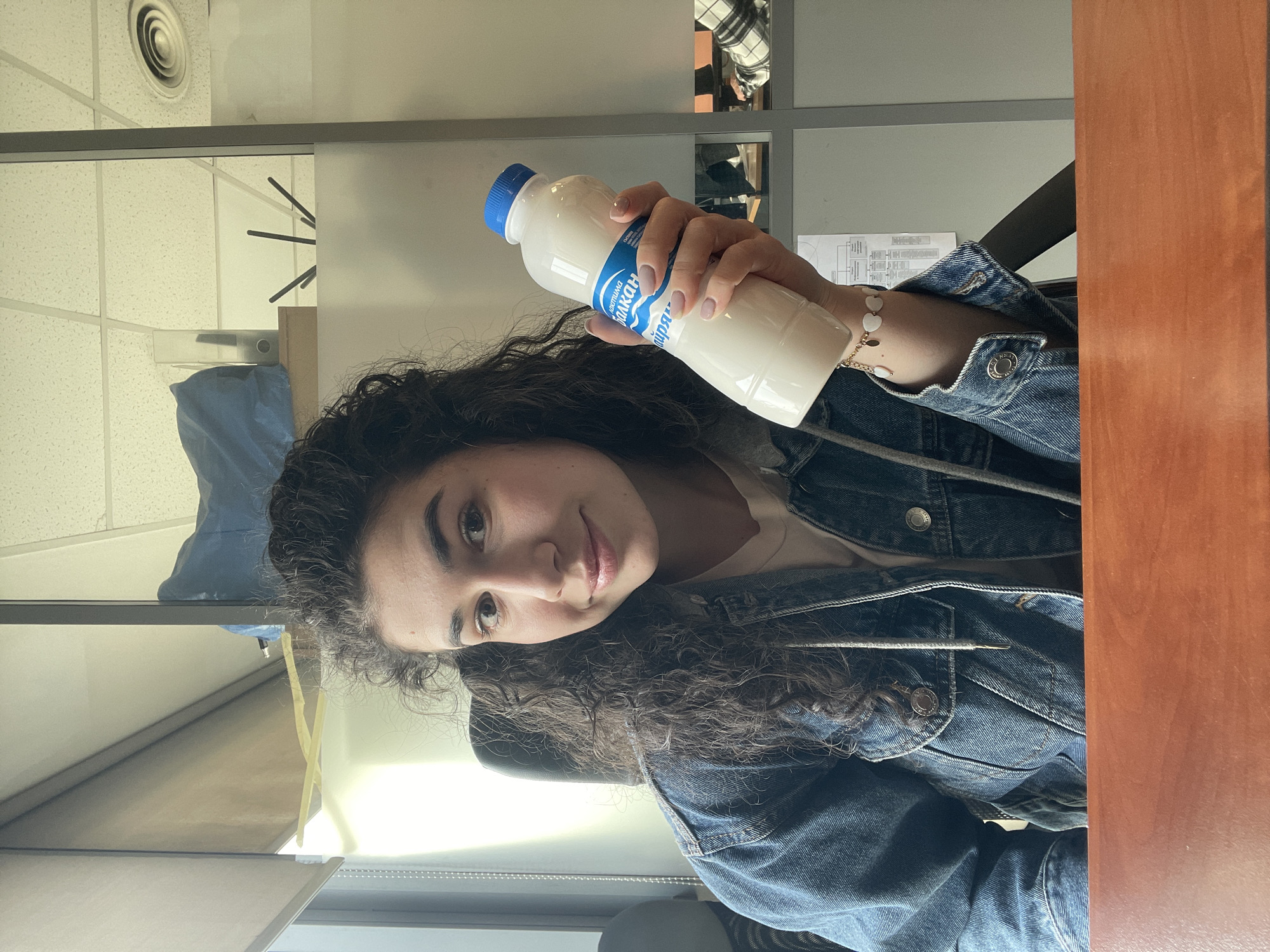 A person with dark curly hair, wearing a denim jacket and a bracelet, is holding a white plastic bottle with a blue label and cap. The label appears to be in Cyrillic script. The setting appears to be an office, indicated by the presence of desk panels, a ceiling with air vents, and office equipment. The desk is wooden. The location is potentially in Sofia, Bulgaria, based on the Cyrillic lettering on the bottle label.
