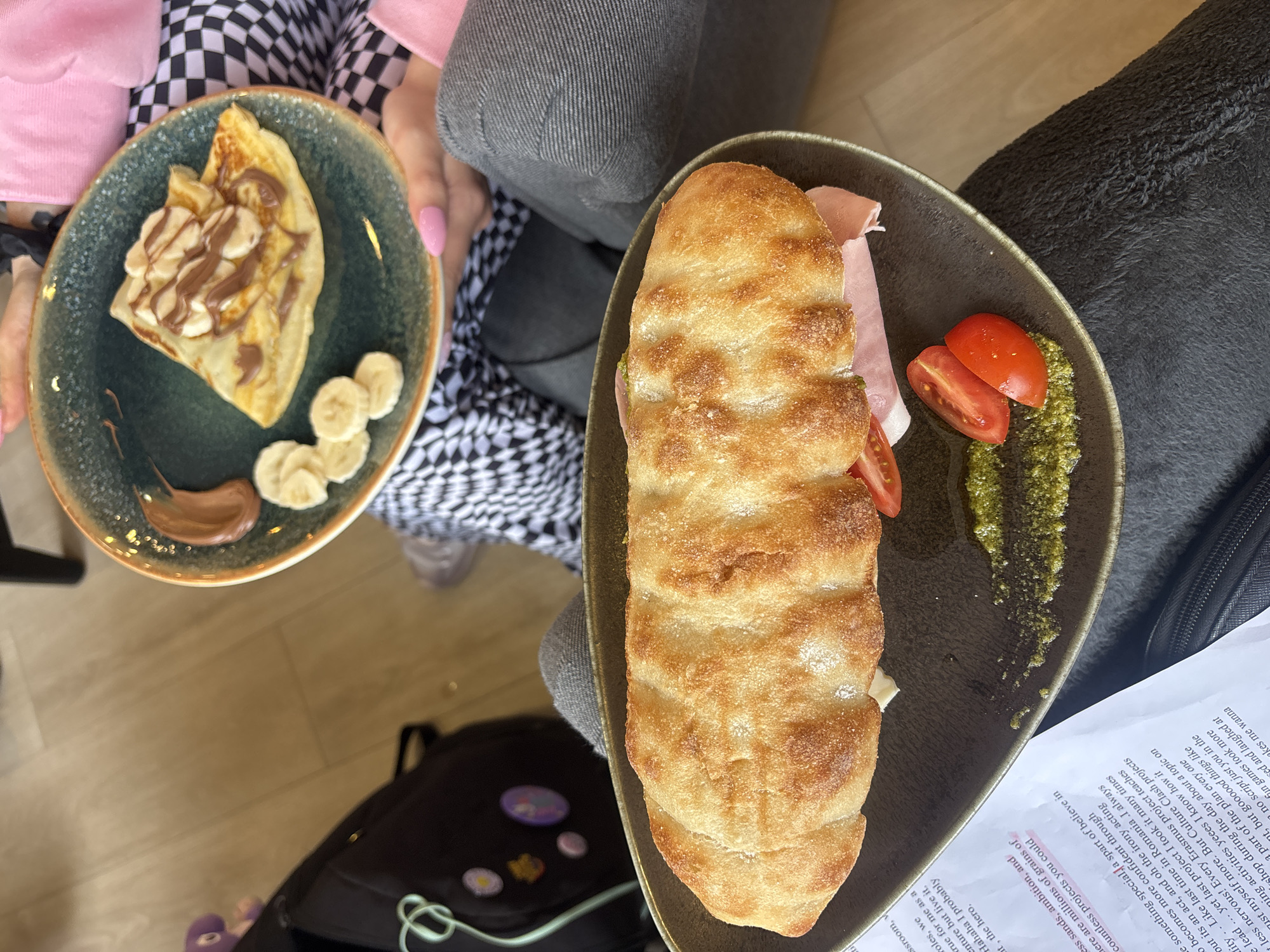 The image shows two plates of food. On the left, a person with a pink sweater and a black and white checkered garment is holding a plate with a folded crepe, covered with chocolate and slices of banana. A dollop of chocolate sauce sits on the side of the plate. On the right, a plate contains a sandwich with bread, ham, and tomatoes, accompanied by pesto. Part of a book lies next to the right plate, with visible text. A black bag is near the plates. The location is not specified.