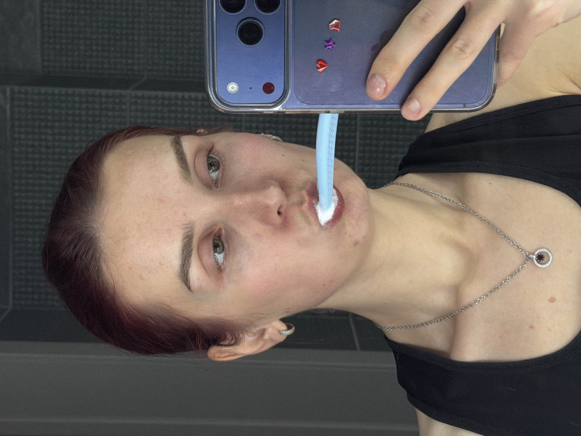 A rotated, close-up selfie captures a young woman, likely in her late teens or early twenties, brushing her teeth. She has reddish-brown hair, fair skin, and green or hazel eyes, and is looking directly towards the phone's lens. A light blue electric toothbrush, with white toothpaste foam around the bristles, is visible inside her mouth.
She holds a purple-colored smartphone, possibly an iPhone, in a clear case adorned with small, colorful star and heart-shaped glitter stickers. The phone's main camera array is clearly visible, reflecting its use for the self-portrait. She wears a simple black tank top or sports bra and a silver chain necklace with a circular pendant featuring a reddish-brown gemstone surrounded by smaller clear stones. A silver hoop earring is visible in her left ear.
The background suggests an indoor setting, likely a bathroom, with dark tiled walls on one side and a plain, darker surface on the other. The image is oriented horizontally, with the woman's head facing what would typically be the top of a vertically-oriented image. The activity takes place in Chaikovsky, Russia.