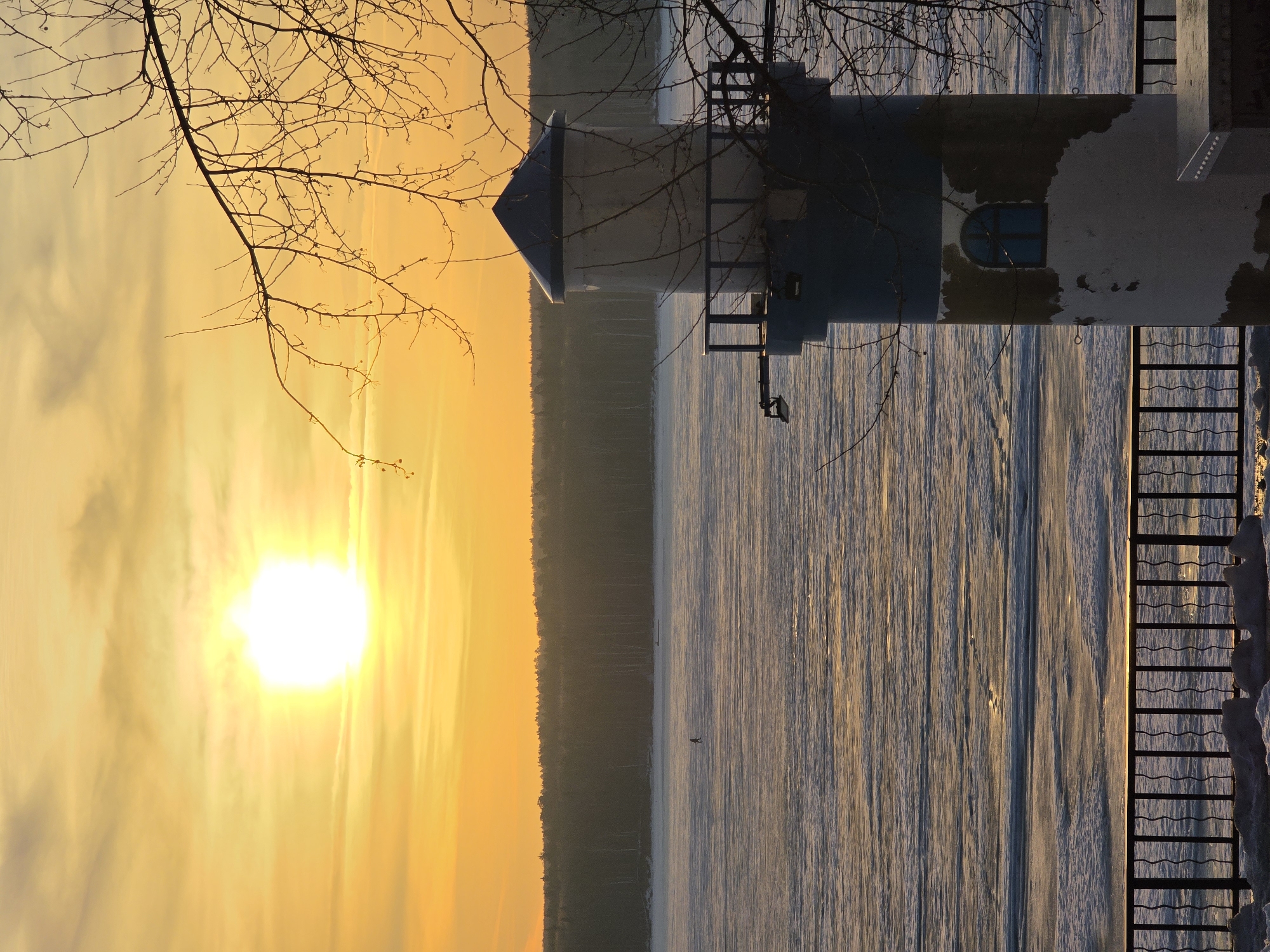 The image depicts a winter scene in Chaikovsky, Russia, at sunset or sunrise. A partially frozen body of water, possibly the Kama River, is visible with ice formations and a dark figure that could be a person on the ice. A forested area borders the water. A building with a light-colored tower and a blue-framed window is located on the right side of the image, near a railing partially covered in snow. Bare tree branches are in the foreground. The sun is low on the horizon, casting a golden glow across the sky.