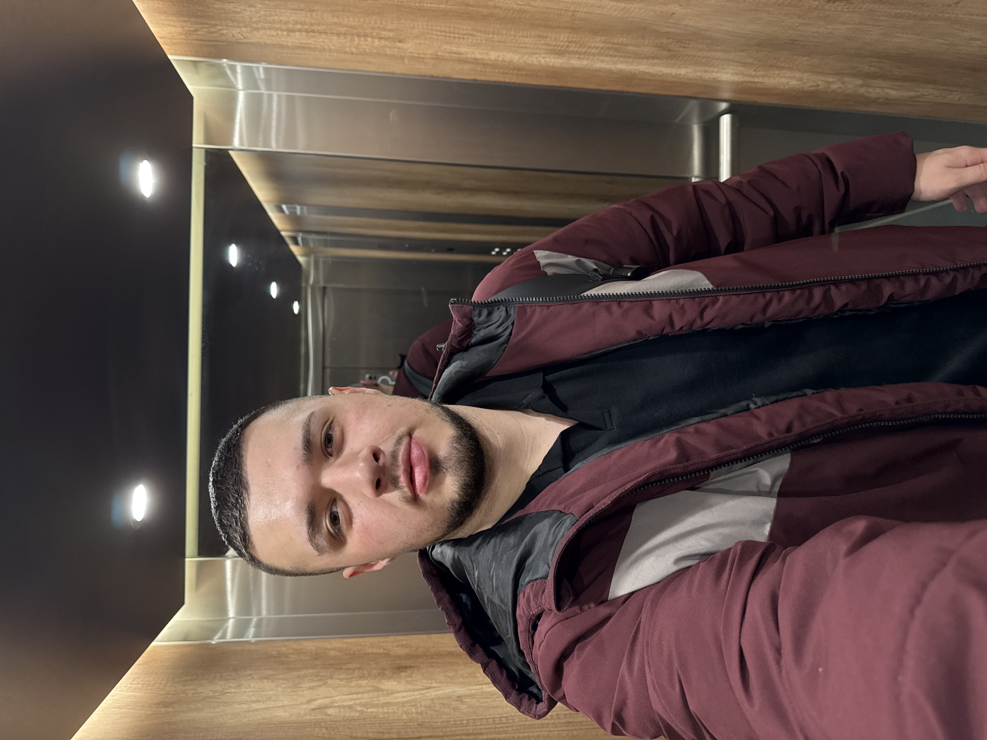 A medium, close-up shot, slightly rotated counterclockwise, shows a man with a short haircut and trimmed beard in an elevator. The elevator features wooden panels on the walls, a metallic back, and small recessed lights on the left side. The man is wearing a black shirt and a burgundy jacket with gray accents. He is looking directly at the camera. The location is not explicitly identifiable as Sofia, Bulgaria, from this image alone.