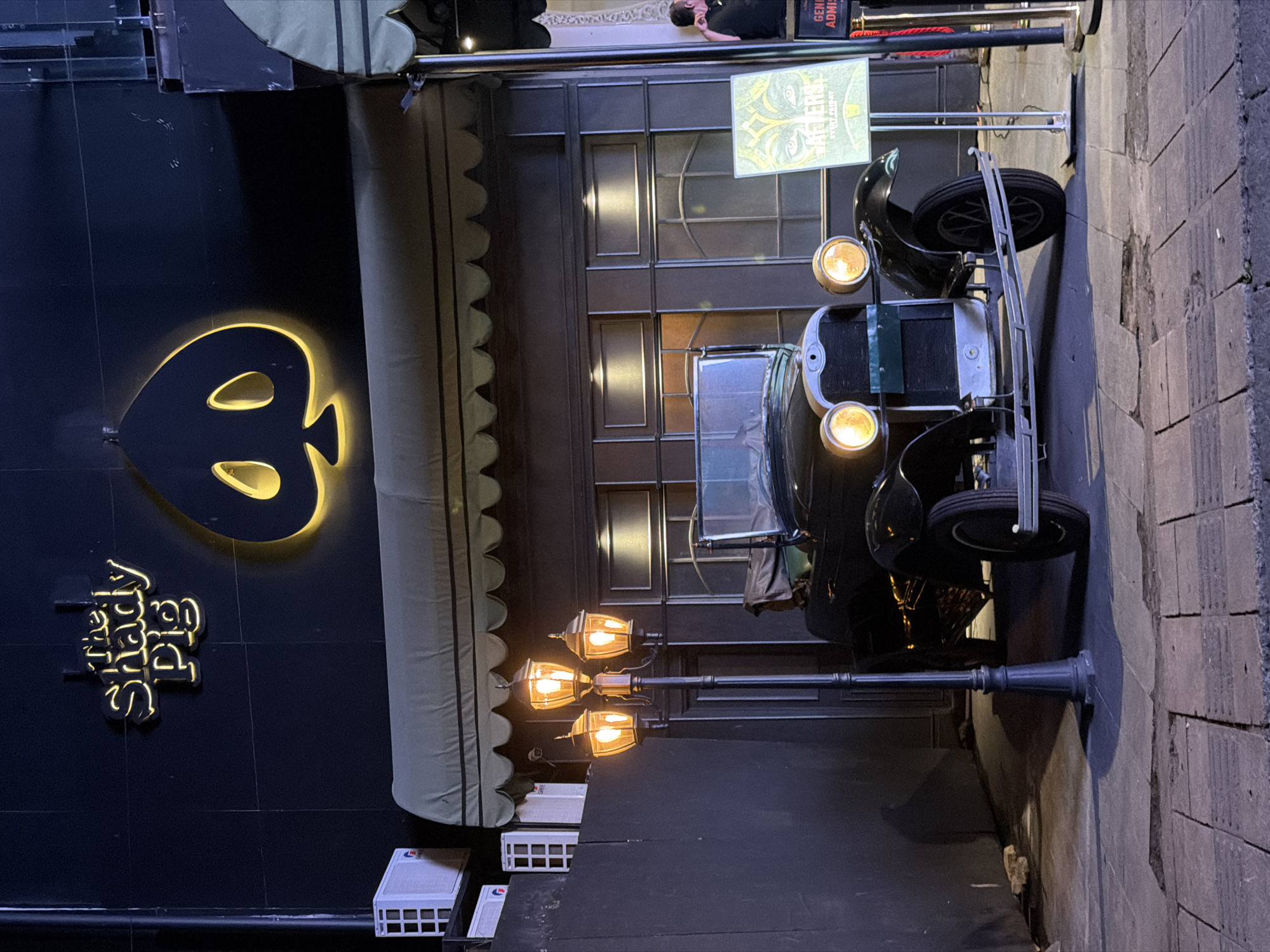 This media file captures a nighttime street scene in North Kuta, Indonesia, showcasing the facade of a building and a vintage car. The image is taken at a low angle, emphasizing the illuminated elements against the dark setting.
The main subject on the left is a building with a dark, possibly black, exterior. A prominent sign, "The Shady Pig," is illuminated in glowing yellow script, accompanied by a large, glowing yellow symbol resembling a stylized spade or a pig's head/skull. Above the entrance, there's a light-colored awning with scalloped edges. Further up the building, a large display poster is visible with abstract artwork and text that appears to read "BREKER" (or similar) and "EVENT TICKET," along with "GENR ADMISS" (General Admission). A person is partially visible near this sign, lying or leaning against a surface.
To the right, a classic black vintage car, possibly an antique Ford Model T, is parked on a paved surface, likely a sidewalk or street. Its two large, circular headlights are brightly illuminated, and a spare tire is mounted on its side or rear. In front of the car, a tall lamppost with three ornate, glowing lanterns adds to the nostalgic ambiance. The paved ground shows distinct block patterns, resembling cobblestones. At the base of the building, several white rectangular boxes, possibly air conditioning units or electrical components, are visible.
The overall impression is that of a themed establishment, likely a bar or club given the name "The Shady Pig" and the event sign, creating a speakeasy-like atmosphere with the vintage car and old-fashioned lighting. The time of day is clearly night, as indicated by the illuminated signs, car headlights, and streetlights.