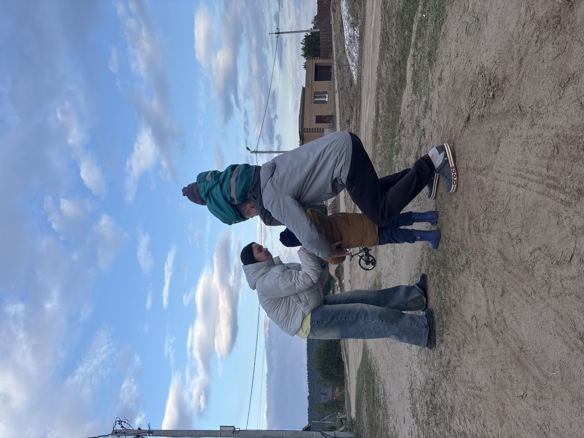 The image, set in Chaikovsky, Russia, captures a playful outdoor scene featuring three individuals forming a human pyramid on a dirt path. At the bottom, a woman wears a light grey puffy jacket, blue jeans, and a dark beanie, supporting the person above her at their waist. Above her, a male individual, dressed in a light grey jacket, black pants, and dark grey Crocs, is bent forward in a plank-like pose. The word "crocs" is visible on the side of his shoe. Balanced on his back is a small child, clad in a teal/green padded jacket and a dark beanie. Partially visible behind this central group, a fourth individual, possibly a child, wears a brown jacket and blue rubber boots and is near a small bicycle wheel.
The ground is a dirt road or path with visible tire tracks and sparse patches of grass. In the background, there are modest houses, fences, and distant trees, characteristic of a semi-rural or residential area. The sky is bright blue with scattered white, wispy clouds, indicating clear weather. The soft, diffused lighting suggests the time of day is likely late afternoon or early evening. The overall scene conveys a sense of casual family activity.