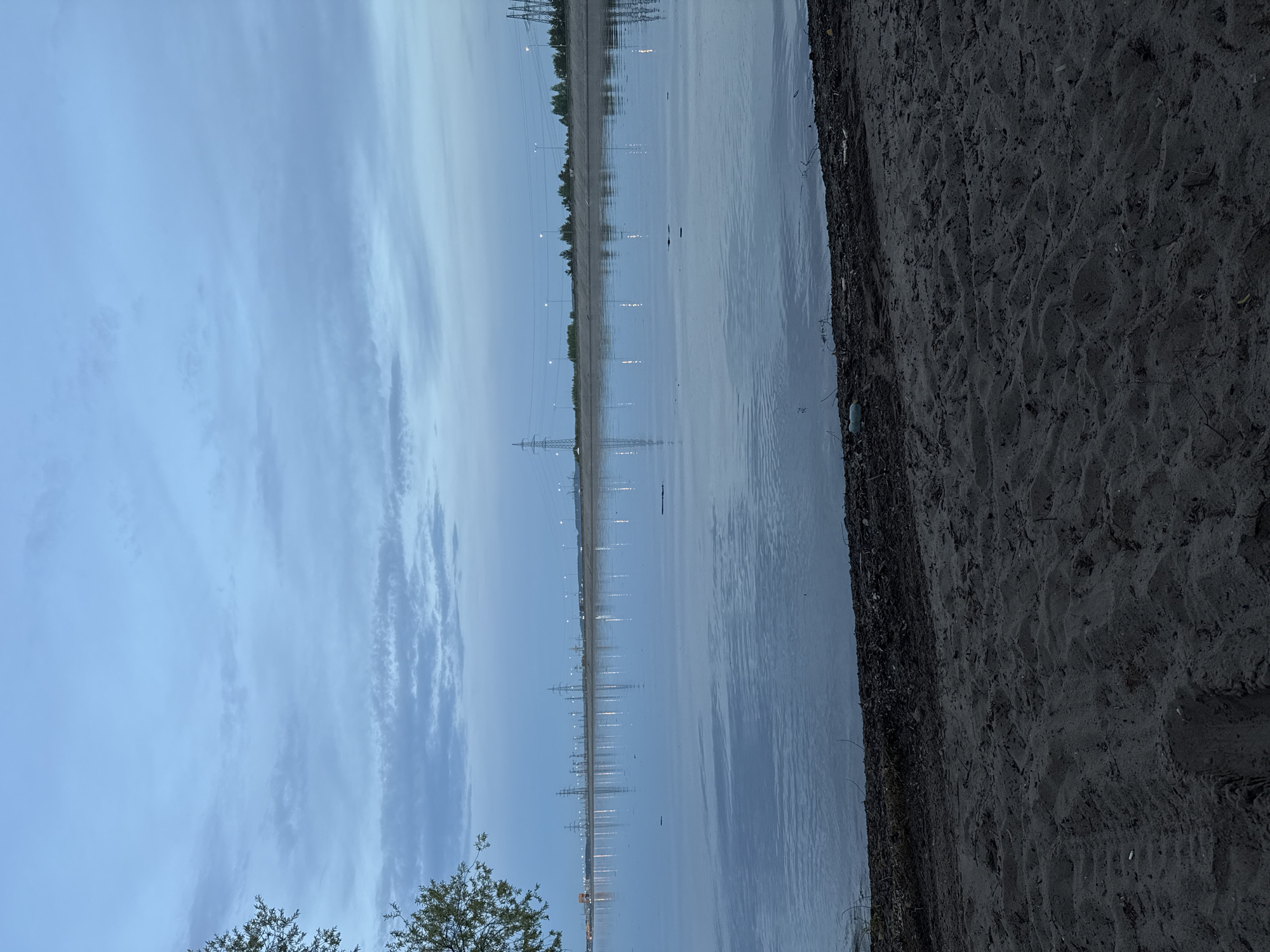 The image depicts a waterside scene in Noviy, Russia. The foreground features a dark sand beach leading to the water's edge. In the middle ground, there is a body of water reflecting the sky and lights along the opposite shore. The far shore is lined with trees, streetlights, and electrical transmission towers that extend along the horizon. The sky is partly cloudy. The overall scene is twilight. No people are visible.