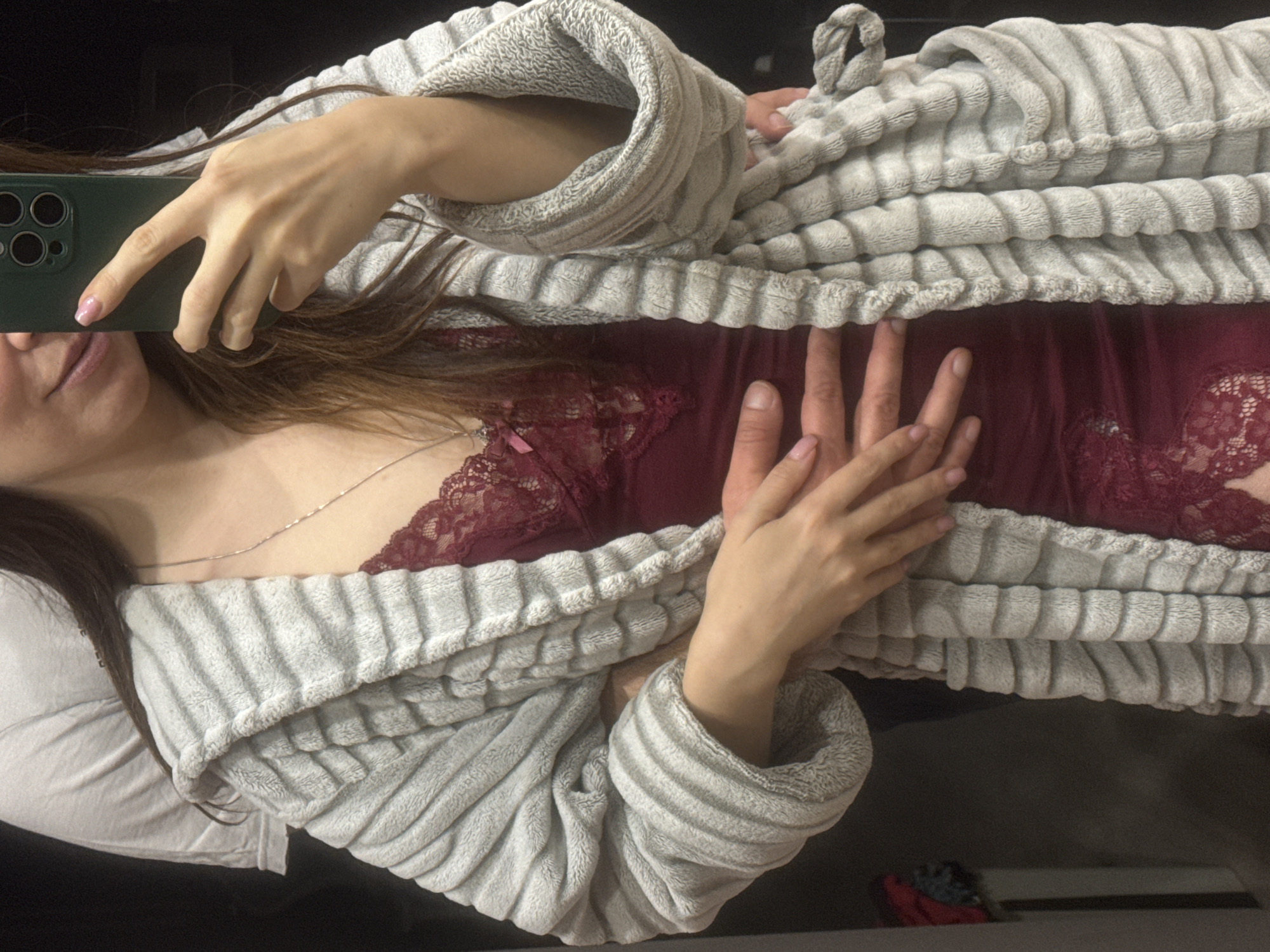 Inside a private residence, city of Unknown, Unknown.
A close-up, waist-up mirror selfie depicts two individuals. A female, positioned in the foreground, faces the camera. She has visible brown hair and wears a grey, plush, horizontally ribbed bathrobe over a burgundy lace-trimmed camisole. A thin silver necklace is visible on her chest. Her lower face is partially visible, showing a slight smile. Her right hand, with pink fingernail polish, holds a dark green smartphone with a visible multi-lens camera module, positioned to capture the image. Her left hand is placed over the left hand of a male individual, whose arm is wrapped around her waist. The male is partially visible behind the female, wearing a light grey short-sleeved t-shirt. His left hand rests on the female's abdomen. The background is dark and out of focus, with a partial view of a storage unit or wardrobe containing red and blue fabric items visible on the far left.