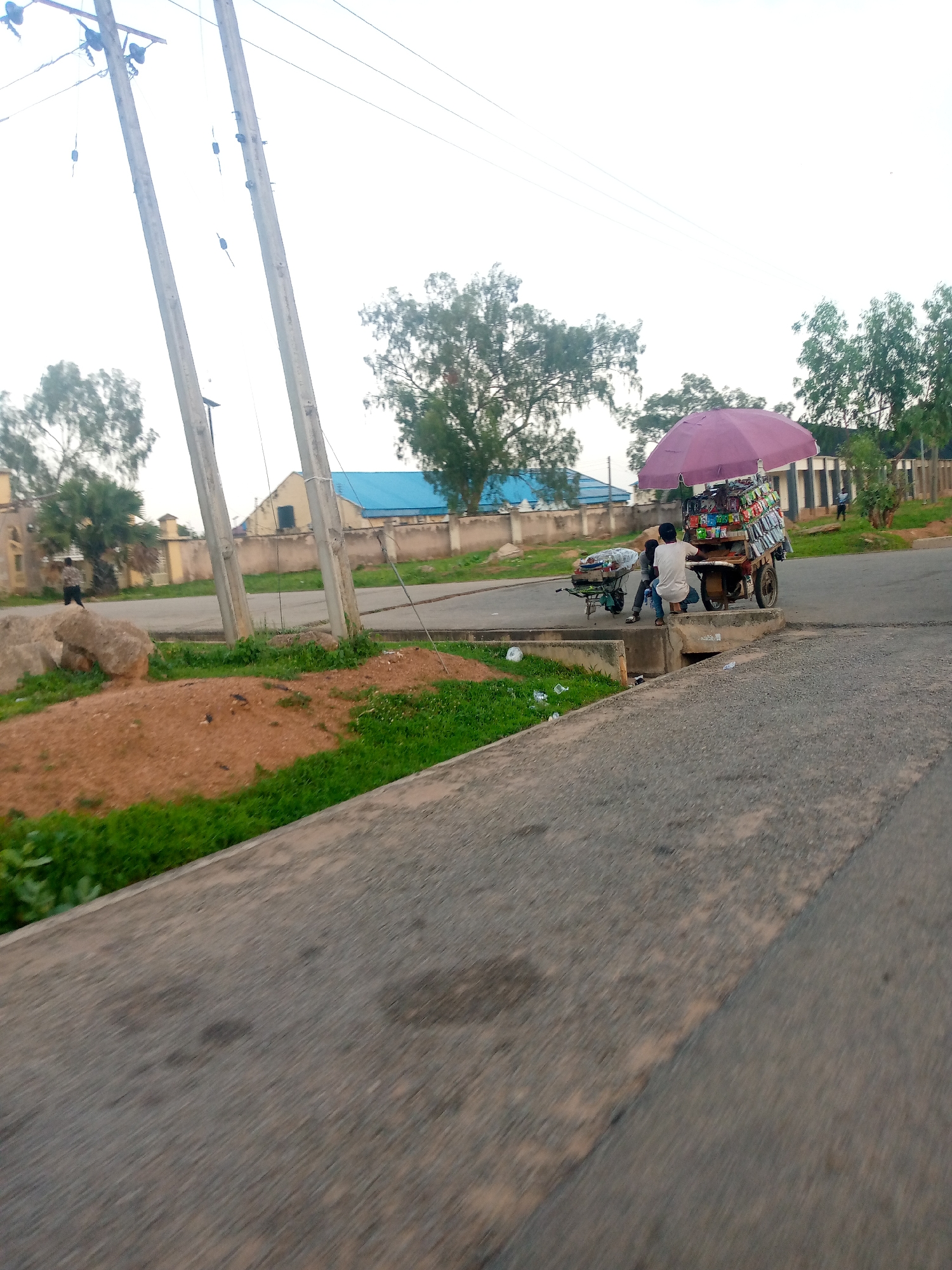 The image depicts a street scene in Jalingo, Nigeria, likely on an overcast day. In the foreground, a paved road is visible, with a dirt shoulder on the left side covered in dry grass and some scattered rocks. Power lines are strung between two tall poles on the left.
Further back, a mobile vending cart laden with various items, possibly phone accessories or snacks, is parked near a drainage ditch. A person sits on the cart, facing away from the camera, while another individual is seated next to them. A large, open umbrella, likely purple, is attached to the cart to provide shade. To the left of the cart, a smaller wheeled cart is also present.
In the background, there are buildings with light-colored walls and blue roofs, and a line of trees. A few other individuals are visible in the distance, one walking along the road and another near the buildings. The overall impression is of a commercial street or area with active street vendors.