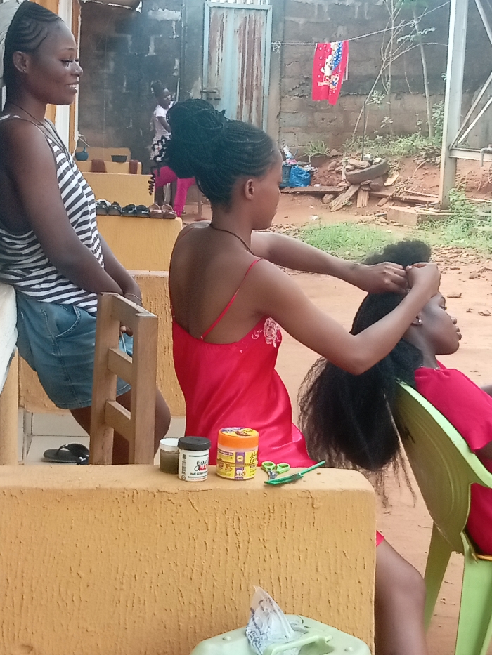 Three women are present. One woman wears a black and white tank top and denim shorts, leaning against a wall. Another woman in a red satin dress braids the hair of a woman seated in a green chair, also wearing red. A row of shoes sits atop the wall. Hair products and scissors are on the wall beside the braider. In the background, a building, a gate, and a clothesline with a red and white cloth are visible. The setting appears to be outdoors.