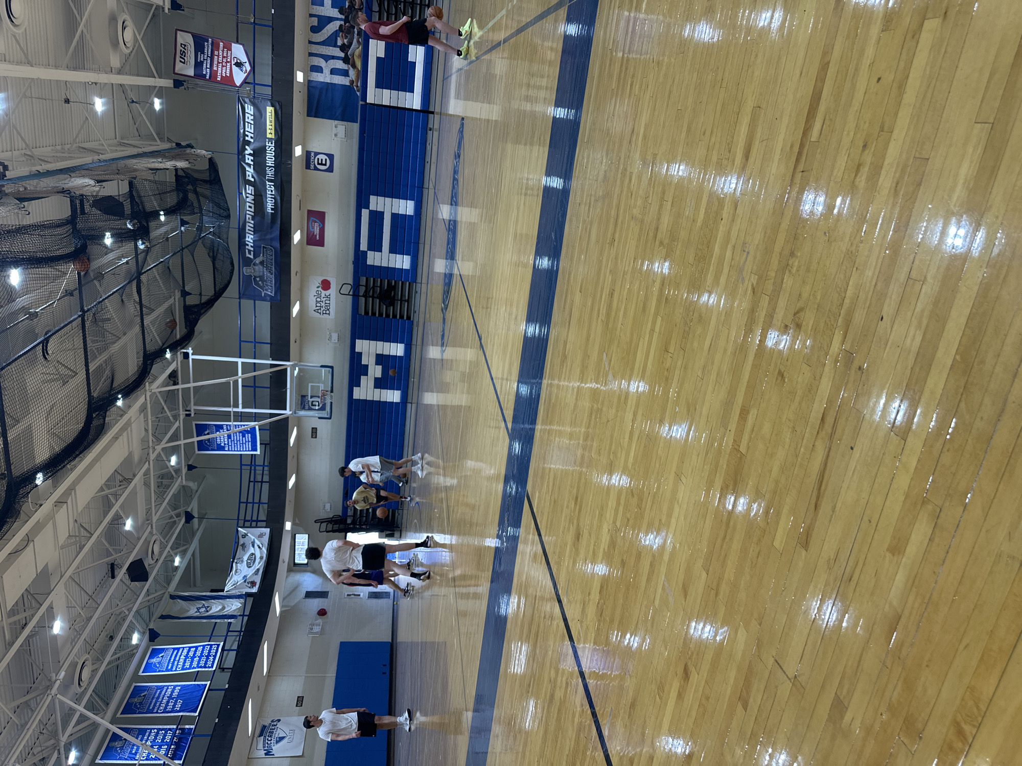 This media file captures an indoor basketball court, likely a college or university gymnasium, bathed in bright artificial light that reflects off the highly polished wooden floor. Several young men, dressed in athletic t-shirts and shorts, are present on the court, appearing to be engaged in a basketball practice or warm-up session. Some are standing, while others are in bent-over postures, possibly stretching or preparing. Basketballs are visible on the court.
The gymnasium features blue bleachers along one side, with large white lettering that reads "MACCABEES." A protective black netting is seen near one of the basketball hoops. Numerous banners hang from the high ceiling, celebrating team achievements and displaying motivational slogans. Visible text includes "CHAMPIONS PLAY HERE," "PROTECT THIS HOUSE" with an Under Armour logo, and "MACCABEES UNITE." Banners list championship years such as "2014, 2015," "2016, 2017," "2018-2019," and "2017-2018, 2019-2020, 2021-2022, 2024-2025." A "SECTION E" sign is also visible. An "Apple Bank" logo is prominently displayed on the bleachers. One banner, distinct from the basketball championships, announces the "USA Hockey Collegiate Division III National Championship, April 17-21, 2024, Auburn, Maine," suggesting a multi-sport facility or a broader institutional affiliation. Based on the "MACCABEES" team name and the provided location context, this facility is likely in New York, United States.
