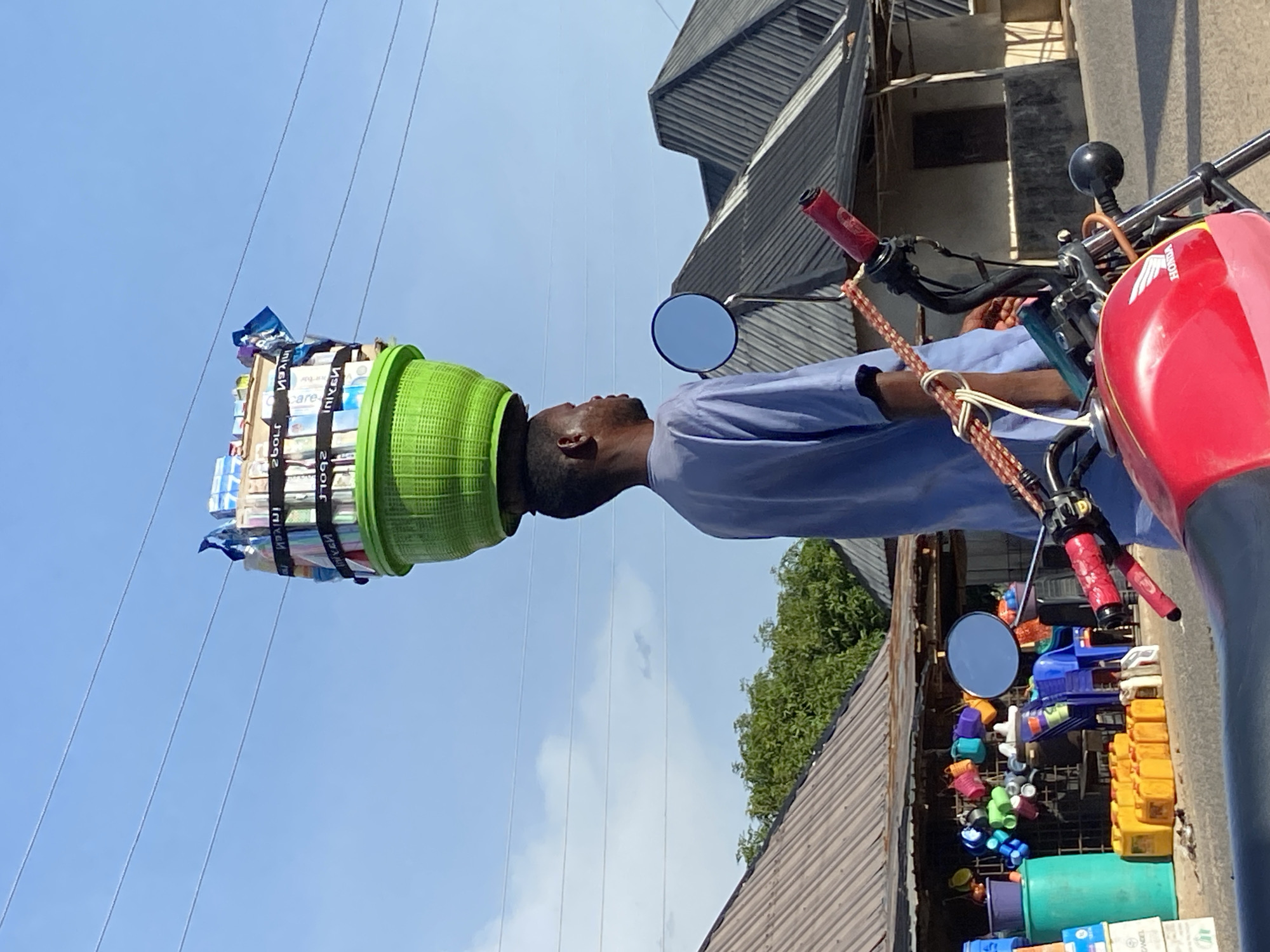 In Mararaba, Nigeria, a man balances a green plastic basket filled with goods on his head. The basket, secured with black straps, contains packaged items. The man is dressed in a light blue shirt. A red Honda motorcycle is parked nearby, laden with colorful plastic containers. Overhead electrical wires traverse the scene, set against a clear blue sky. Buildings with corrugated metal roofs line the street.