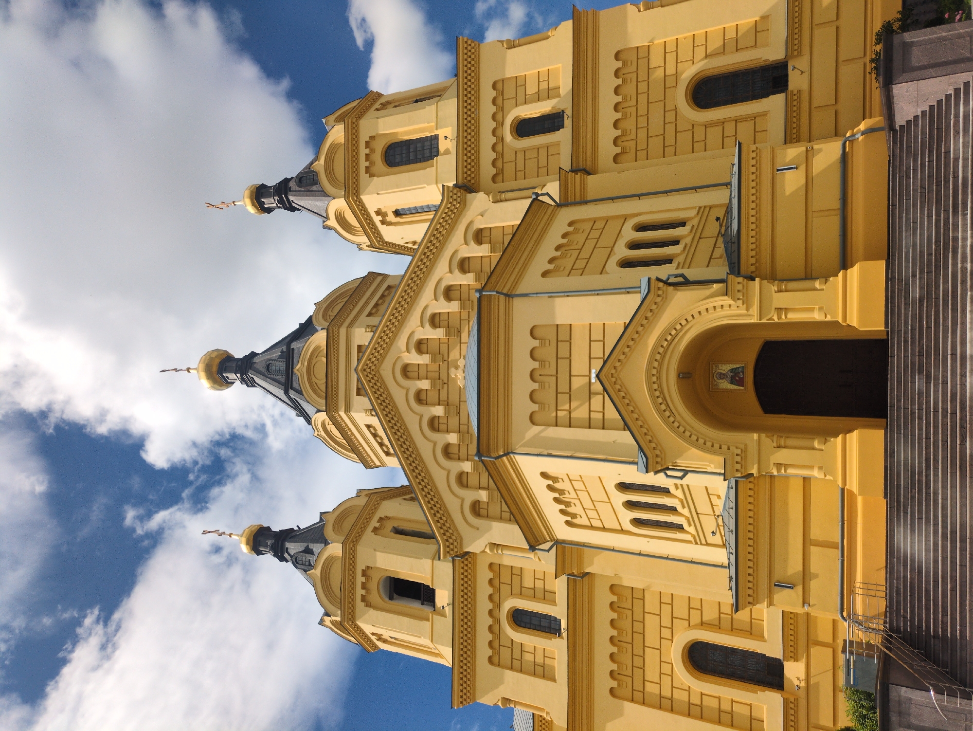 This is a low-angle, exterior, full shot of the Alexander Nevsky Cathedral in Nizhny Novgorod, Russia. The cathedral is a large, yellow building with a dark, conical roof topped with a golden dome and cross. It has multiple domes, arches, and intricate architectural details. The sky above is blue with white, fluffy clouds. The foreground shows a wide, stone staircase leading up to the entrance of the cathedral. There are no people visible in the image. The scene appears to be captured on a bright, sunny day.
