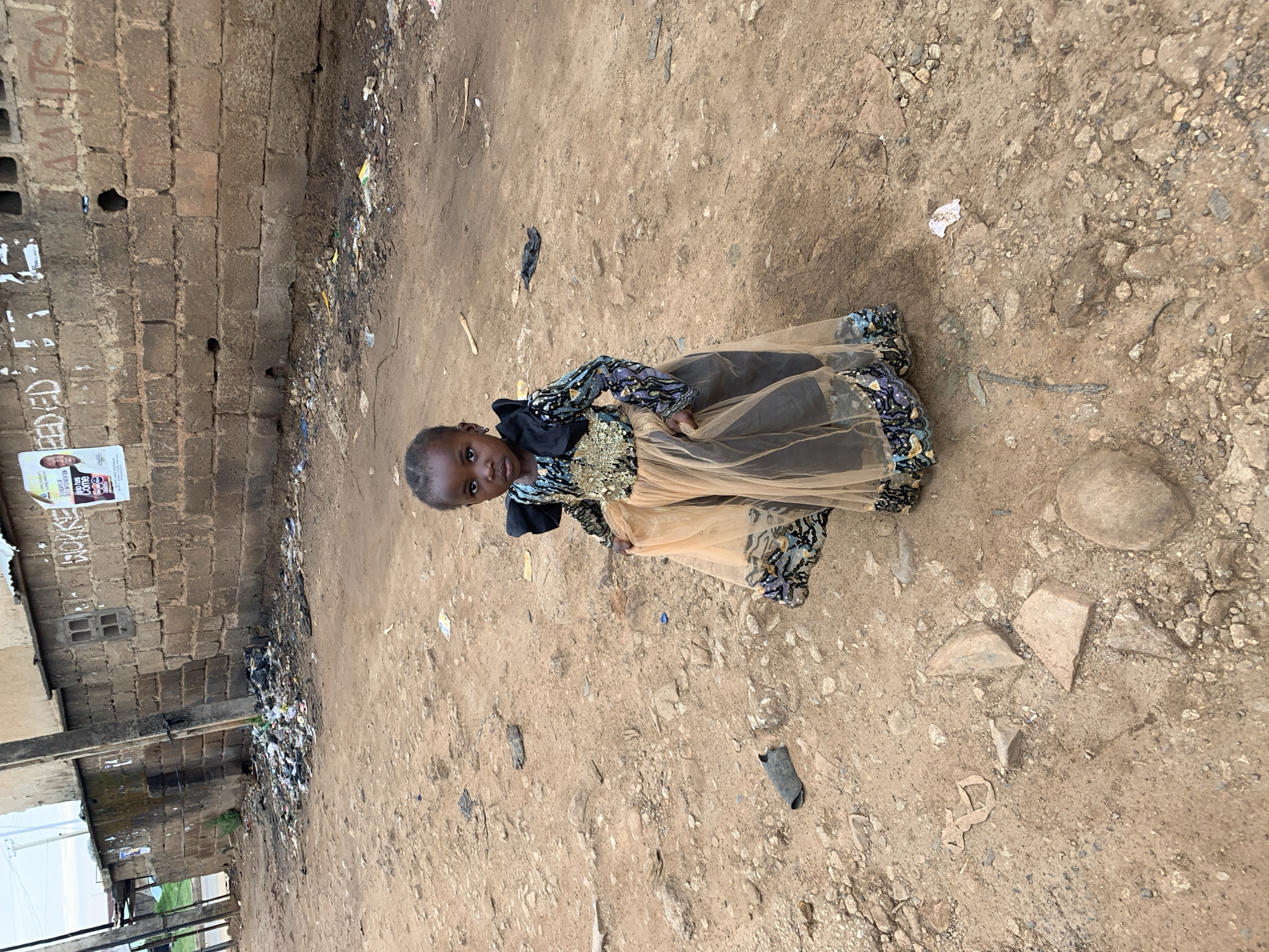 The image depicts a young child, appearing to be a girl, standing on an unpaved dirt road in Rafin Par, Nigeria. The child, with dark skin and short dark hair, faces forward, looking directly at the camera with a neutral expression, wearing small earrings. She is dressed in an ornate, long-sleeved gown featuring a black top adorned with gold and iridescent blue-green sequins or embroidery. The skirt is composed of a sheer, light tan or orange tulle overlaying a darker fabric, with matching sequined patterns on the lower hem. A large black fabric bow or detail is present on her left shoulder.
The ground is dry, light brown earth, uneven and littered with small rocks, pebbles, and scattered debris including plastic, fabric scraps, and pieces of wood. In the background, a weathered brick wall extends horizontally, exhibiting dark stains, several rectangular openings or vents, and faded white painted text, including partial words like "REEDFD" and "VARITSA". Attached to the wall are several posters, one prominently displaying a Caucasian man's face alongside text such as "5 YEARS OF HIV INTERVENTION" and "Help has Come". Accumulations of mixed refuse, including plastic bags and various debris, are visible along the base of the wall and scattered across the ground. A dark utility pole stands in the mid-ground, extending from the left side of the frame, with another pole visible further back. The sky in the upper left corner appears overcast.