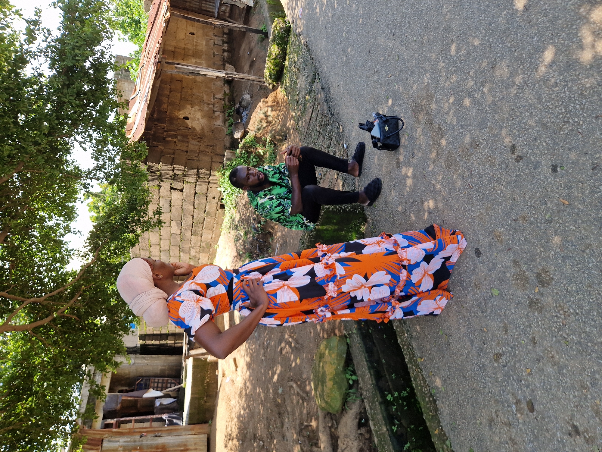 In Kabong, Nigeria, a woman wearing a vibrant orange and blue floral dress and a beige headscarf stands with her hand on her hip, facing away from the camera. Beside her, a man in a green patterned shirt and black pants sits on a concrete block, looking down and holding his hands clasped. A black handbag rests on the asphalt nearby. The background consists of rustic, weathered brick buildings and an overhead tree with lush green foliage, creating a scene of everyday life in this Nigerian locality.