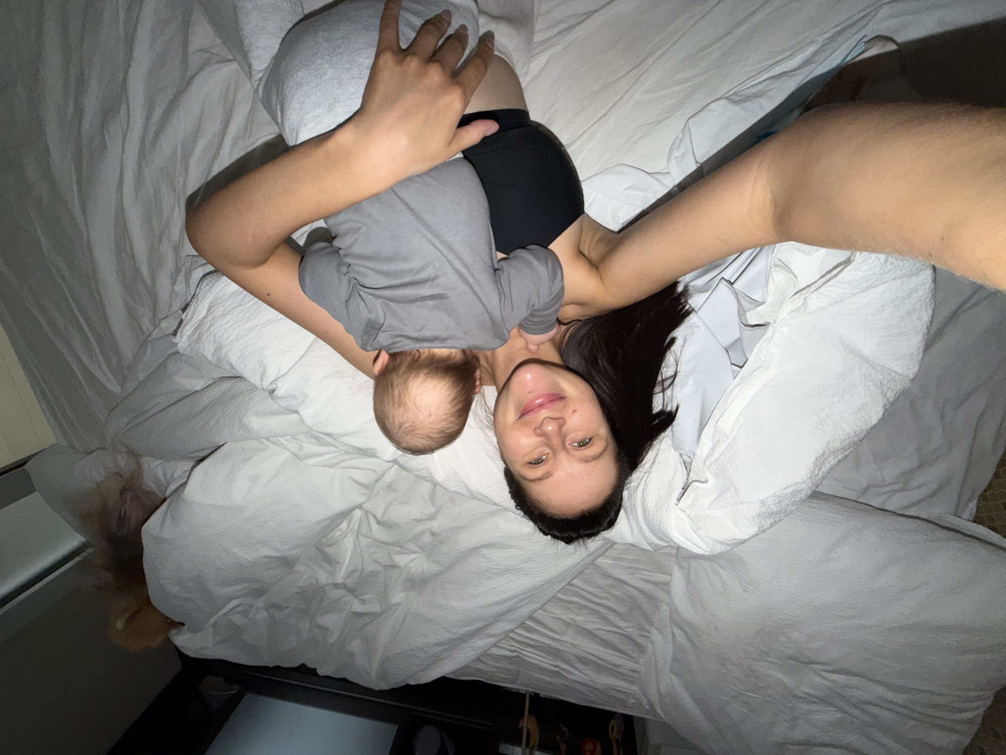 An overhead, intimate selfie captures a woman and a baby lying together on a bed with white, textured bedding, in what appears to be a dimly lit room. The woman, with long dark hair, is smiling gently at the camera. She is wearing a black garment, possibly a top or shorts, and has one arm extended, likely holding the camera.
Cradled against her, the baby is dressed in a grey long-sleeved top and grey pants or a diaper cover. The back of the baby's head is visible as it lies with its head near the woman's chest, seemingly peaceful. One of the baby's arms is bent, resting near the woman's side.
On the lower left side of the bed, partially visible beneath the bedding, is the head of a light-colored dog, adding to the cozy and domestic scene. The soft, low lighting suggests either nighttime or a subdued, cozy atmosphere indoors. The overall image conveys a moment of calm and affection among the family members, likely taken in Khimki, Russia. No visible text is present.