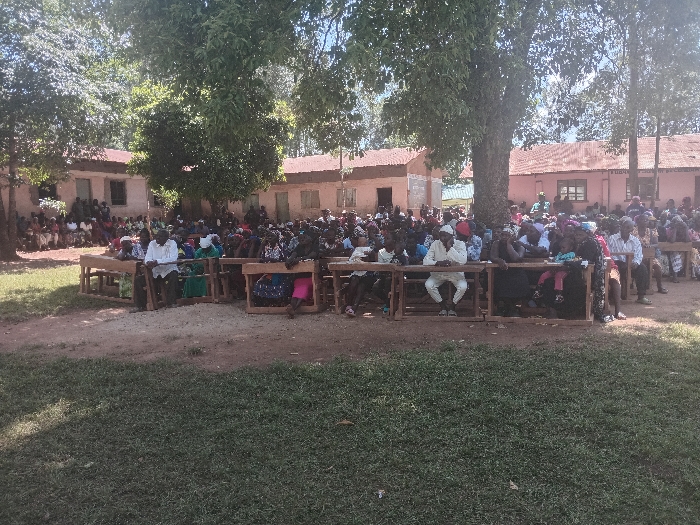 An outdoor public gathering is depicted in Idakho South ward, Kenya. Approximately 150-200 individuals, primarily adults, are assembled in a dirt clearing featuring patches of grass and shaded by multiple large trees. Numerous people are seated on wooden school desks and benches arranged in rows, facing an unseen central point. Additional individuals are visible standing or seated further back near several single-story buildings with light-colored walls and corrugated iron roofs, likely part of an institutional complex. The scene is illuminated by strong natural daylight.