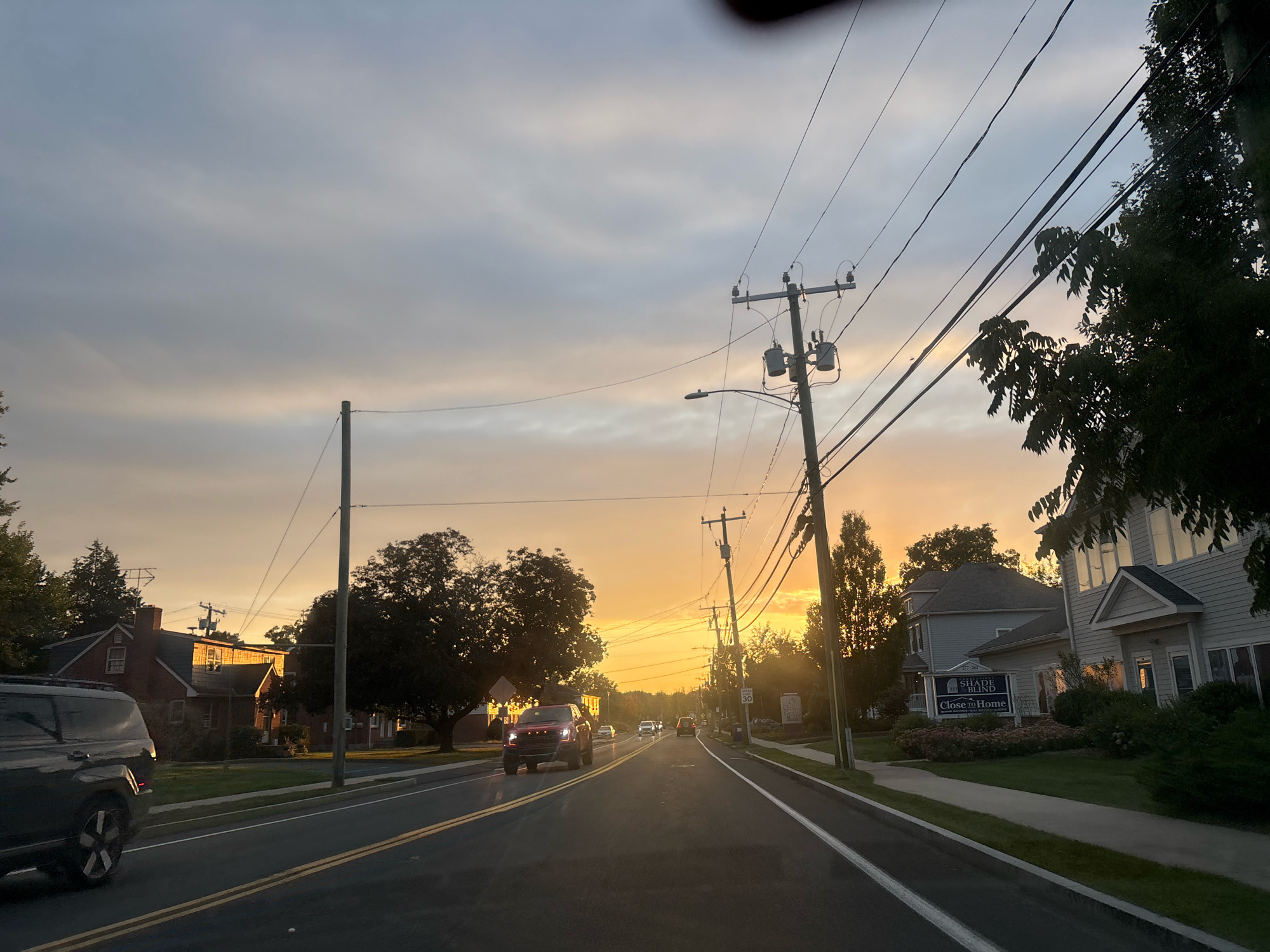The image depicts a street scene in Glastonbury, Connecticut, USA, during a sunset. The sky is filled with a warm gradient of orange and yellow hues, with scattered clouds. A paved road runs through the center of the image, with yellow and white lane markings. Several vehicles are visible on the road, including a dark SUV on the left, a red truck in the middle, and other cars further down.
On the right side of the road, there is a sidewalk and several buildings. One building has a sign that reads "SHADE & BLIND CLOSE TO HOME". Another building to the left appears to be residential, with a brick facade and a chimney. Trees and utility poles with electrical wires are present throughout the scene. The overall impression is a quiet, suburban street at dusk.