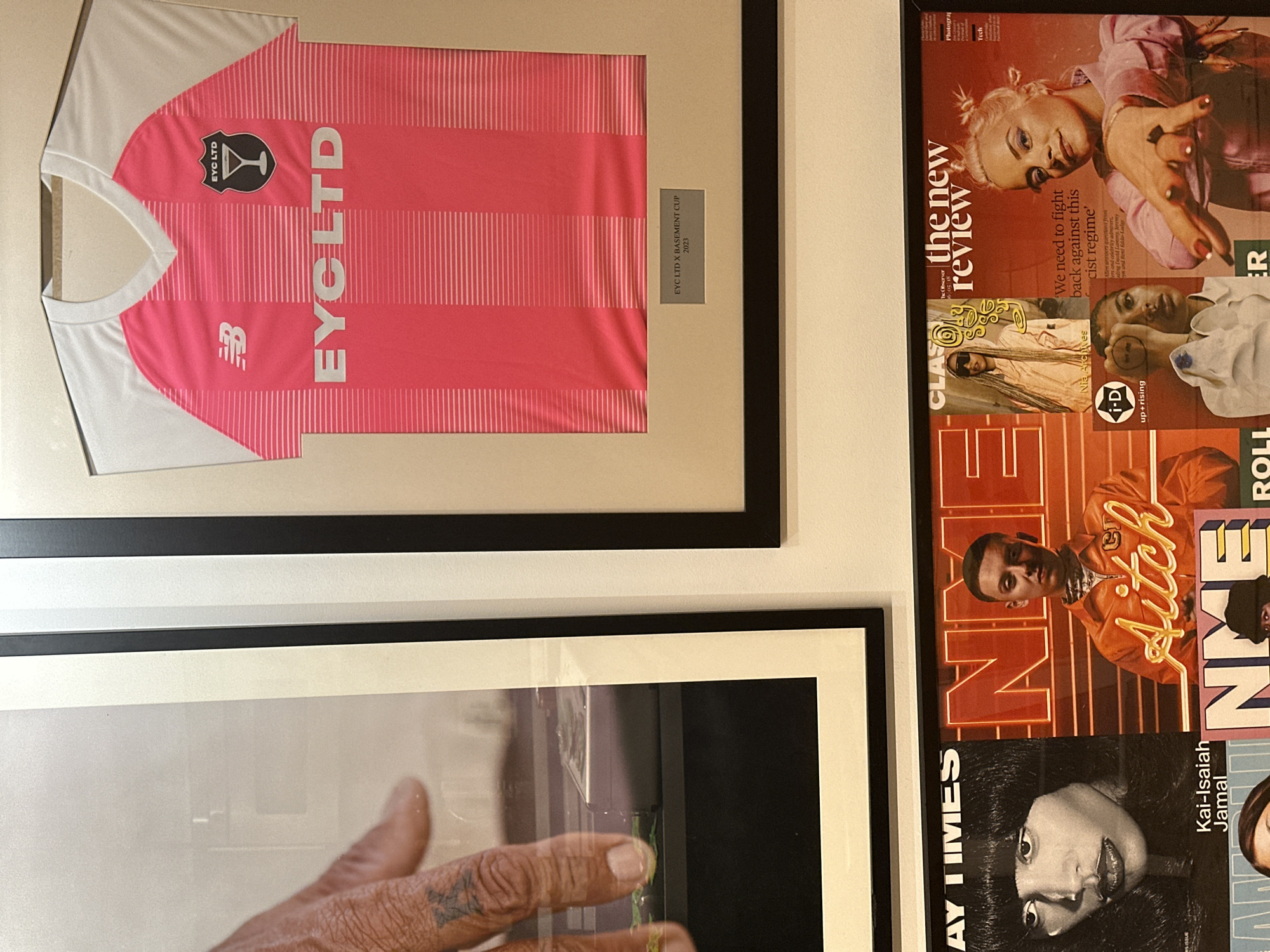 The image displays a wall in an indoor London location, likely a gallery, cultural space, or retail outlet focused on contemporary art, music, and fashion.
The scene features three primary sections:
1. **Top Left:** A framed black-and-white photograph shows a close-up of an individual's left hand, specifically the thumb and index finger. The skin exhibits texture, and a small, dark tattoo resembling an 'X' is visible on the side of the index finger. The hand appears positioned over a dark, reflective surface, possibly an audio device, with a faint green reflection visible. The photograph is encased in a black frame with a white mat.
2. **Top Right:** A framed, folded pink and white football (soccer) jersey is displayed. The jersey features a V-neck, white shoulders, and a pink torso with varying horizontal stripe patterns. A white "NB" (New Balance) logo is on the left chest, and a black shield-shaped logo with "EYC LTD" and a martini glass icon is on the right chest. "EYC LTD" is printed in large white capital letters across the front. Below the jersey, a small plaque reads "EYC LTD X BASEMENT CUP 2023". This item is set within a black frame with a beige mat.
3. **Bottom Section:** A collage of several overlapping magazine covers is arranged.
* A partially visible cover on the far left shows a person identified as "Kai-Isaiah Jamal".
* To its right, an "NME" magazine cover features rapper Aitch in an orange outfit, with his name stylized in orange text.
* Above Aitch, a partially obscured cover mentions "Nia Archives".
* On the far right, "The New Review" cover from The Observer features singer Lily Allen, posed with her hand extended, alongside text stating, "'We need to fight back against this cist regime'".
* Below Lily Allen, an "i-D up+rising" cover shows a person partially obscuring their face with a hand.
The objects are stationary, presented as a curated display on a plain wall, indicative of a creative or commercial environment often found in London's cultural districts. The specific "EYC LTD X BASEMENT CUP 2023" jersey and the featured UK artists on the magazine covers strongly suggest a connection to The Basement's cultural influence, placing the scene within the creative urban landscape of London, United Kingdom.
