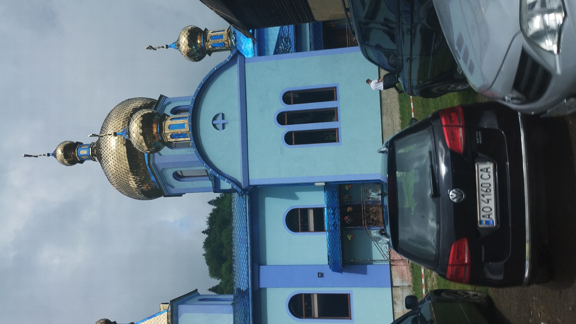 This image depicts the exterior of a bright blue church with golden domes. The building features arched windows and decorative blue trim. Several cars are parked in front of the church. A black Volkswagen is visible in the foreground with a license plate reading "AO 4160 CA". The scene appears to be in Kvasy, Ukraine, indicated by the context. The sky is overcast, suggesting a cloudy day. A person in a white shirt is standing near one of the parked cars. The overall impression is that of a community building in a somewhat rural or suburban setting. The church's vibrant colors stand out against the muted sky.