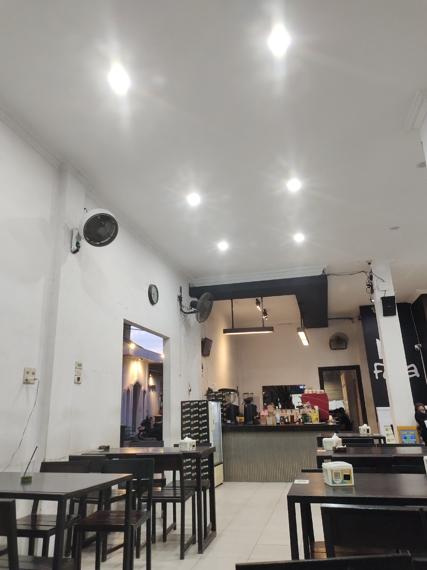 The image depicts the interior of a cafe in Lhoksukon, Indonesia, as indicated by the text on a sign. The perspective is from a low angle, looking upwards and across the space. The cafe features dark wooden tables and chairs arranged in rows. In the foreground, several tables and chairs are visible, with one table in the lower left having a glass with a straw and a green liquid.
The walls are painted white, and the ceiling is white with recessed lighting, some of which are very bright, creating glare. Mounted on the wall is a white oscillating fan. A black analog clock is also visible on the wall. To the right of the clock, another black oscillating fan is mounted lower down. Further to the right, a tall white pillar supports part of the ceiling structure, and on this pillar, a black speaker and some wiring are attached.
A doorway or opening in the wall leads to another part of the building, where a motorcycle is parked and architectural elements with arched windows are visible. Beyond this, the exterior light suggests it might be dusk or early evening.
The main counter area of the cafe is visible in the mid-ground. It has a ribbed metal front and is topped with a dark surface where coffee machines and various bottles of syrups or sauces are placed. A person is partially visible behind the counter, appearing to be working.
On the tables, there are small white boxes, possibly for sugar packets or napkins, and some paper placemats. Signs with text and graphics are visible on some surfaces. In the background, towards the right edge, a person is seated at a table. The floor is tiled with light-colored square tiles. The overall impression is of a modestly appointed, functional cafe.