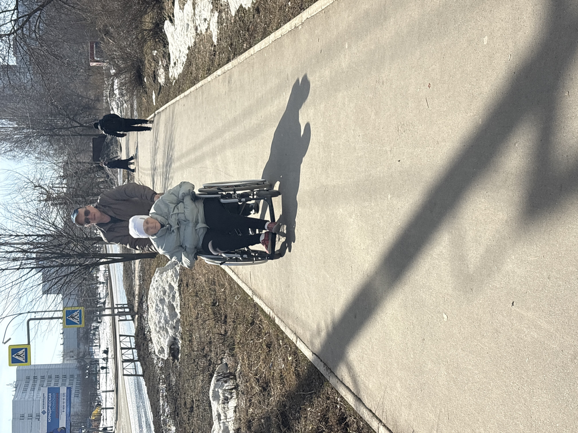 On a sunny day in what appears to be the city of Chaikovsky, Russia, an elderly man pushes a person in a wheelchair along a concrete path. Remnants of snow are visible beside the path, suggesting a late winter or early spring scene. The path has a distinctive shadow cast by the man pushing the wheelchair. Background details include buildings, trees, and street signs. Other pedestrians are further up the path.